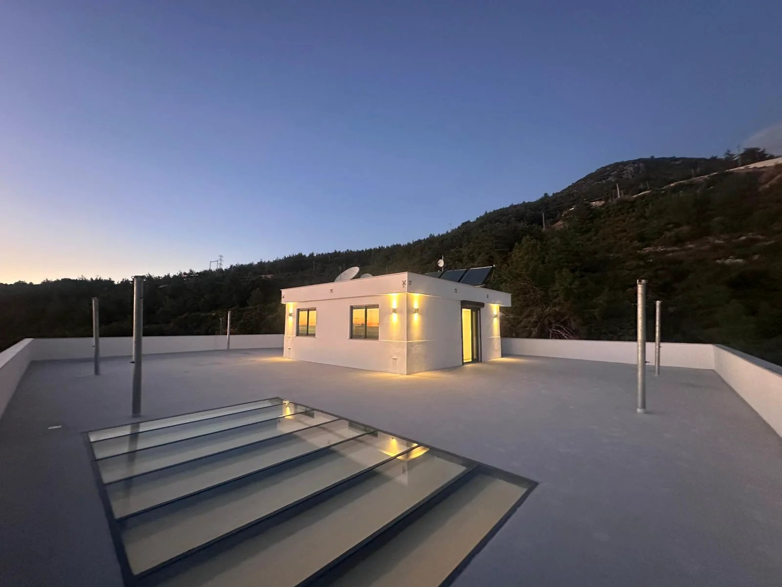 Moderne Dachterrasse mit Bergblick am Hang. Moderne Dachterrasse in der Abenddämmerung mit einer weißen Struktur, warmer Beleuchtung, einem großen Glasdach und Sonnenkollektoren. Bietet einen ruhigen Bergblick unter einem klaren Abendhimmel.