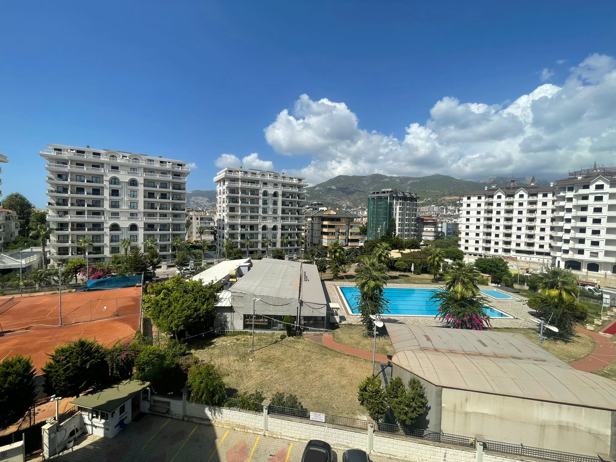 Panoramablick auf moderne Wohnanlage mit Pool und Tennisplatz. Eine Luftaufnahme einer modernen Wohnanlage mit einem großen blauen Swimmingpool, einem Tennisplatz, üppigen Grünflächen, weißen Wohngebäuden und entfernten Bergen unter einem klaren blauen Himmel.
