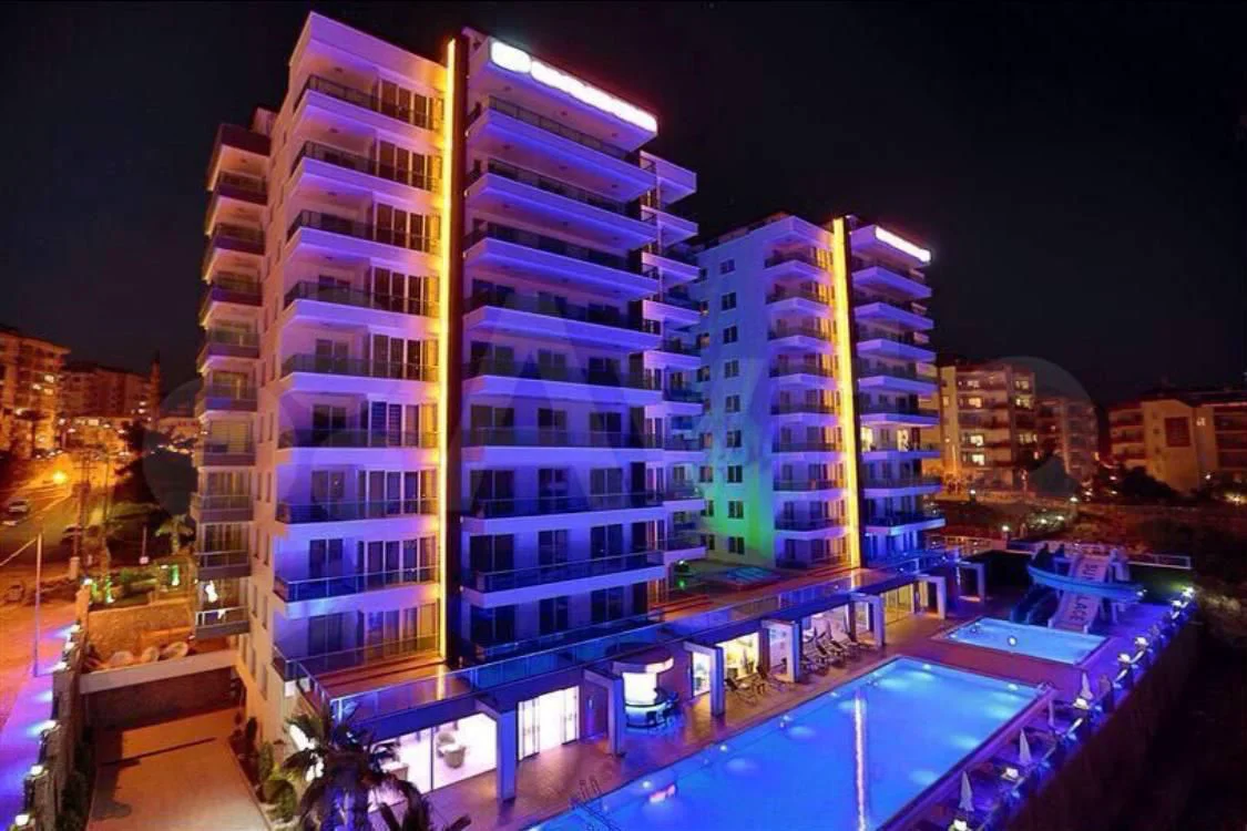 Moderne Wohntürme mit beleuchtetem Poolbereich bei Nacht. Ein moderner Luxus-Apartmentkomplex bei Nacht mit mehrstöckigen Gebäuden mit Balkonen, einem großen, blau beleuchteten Swimmingpool und einem weiteren Pool mit Wasserrutsche, allesamt stimmungsvoll beleuchtet.