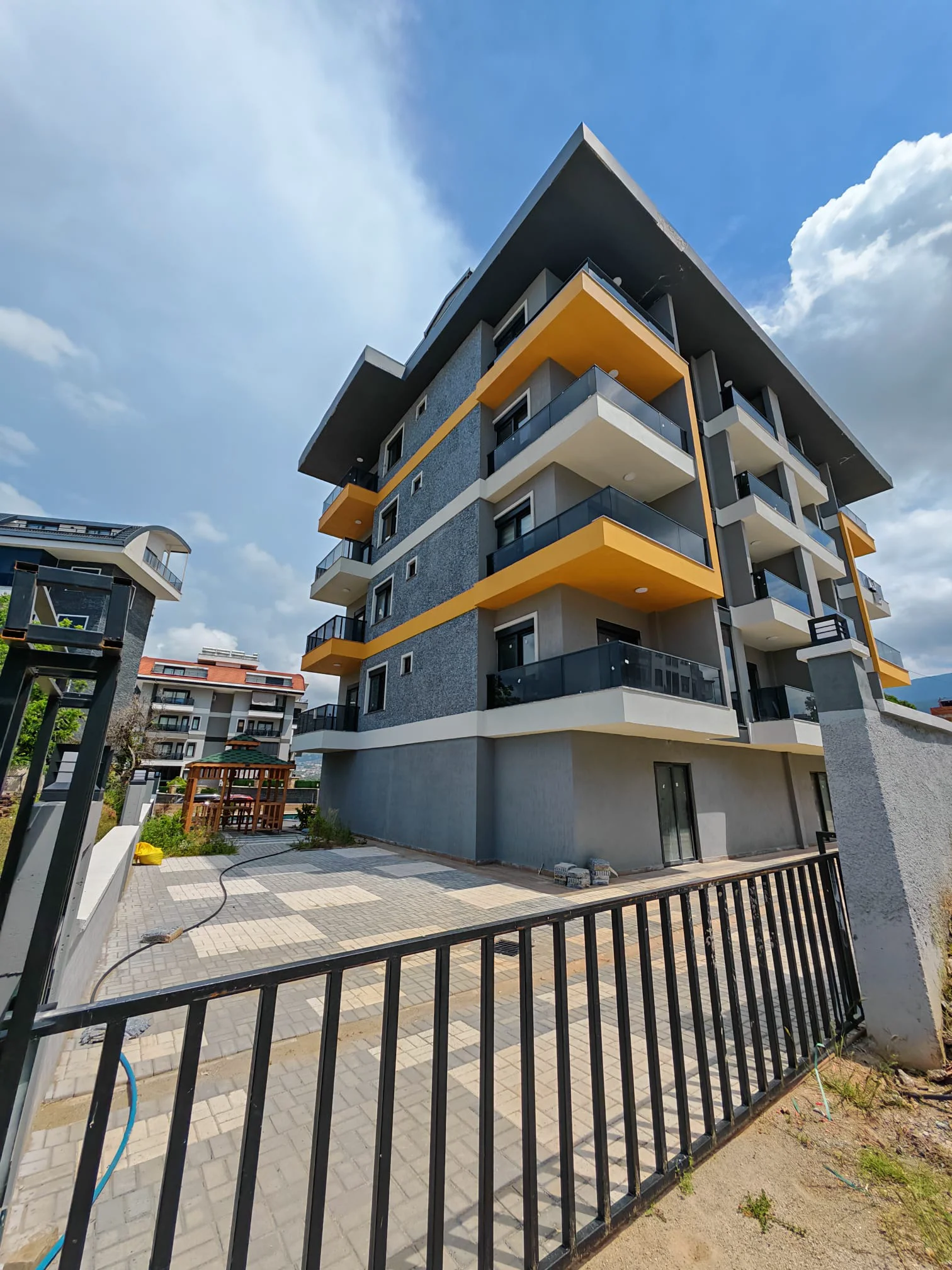 Modernes Apartmentgebäude – Außenansicht mit farbigen Balkonen. Außenansicht eines neu errichteten, modernen Apartmentgebäudes mit grauen und gelben Akzenten, Balkonen mit Glasgeländer und einem gepflasterten Eingangsbereich.