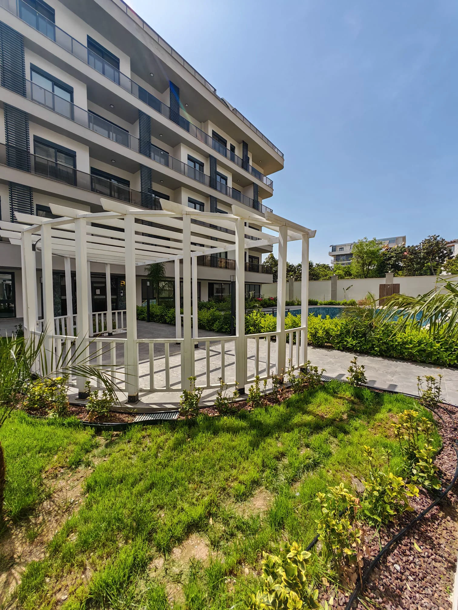 Moderne Außenansicht eines Apartmentgebäudes mit Garten und Pool. Außenansicht eines modernen Apartmentkomplexes mit einer weißen Holzpergola, einem üppigen grünen Gartenbereich und einem blauen Swimmingpool unter einem strahlend sonnigen Himmel.