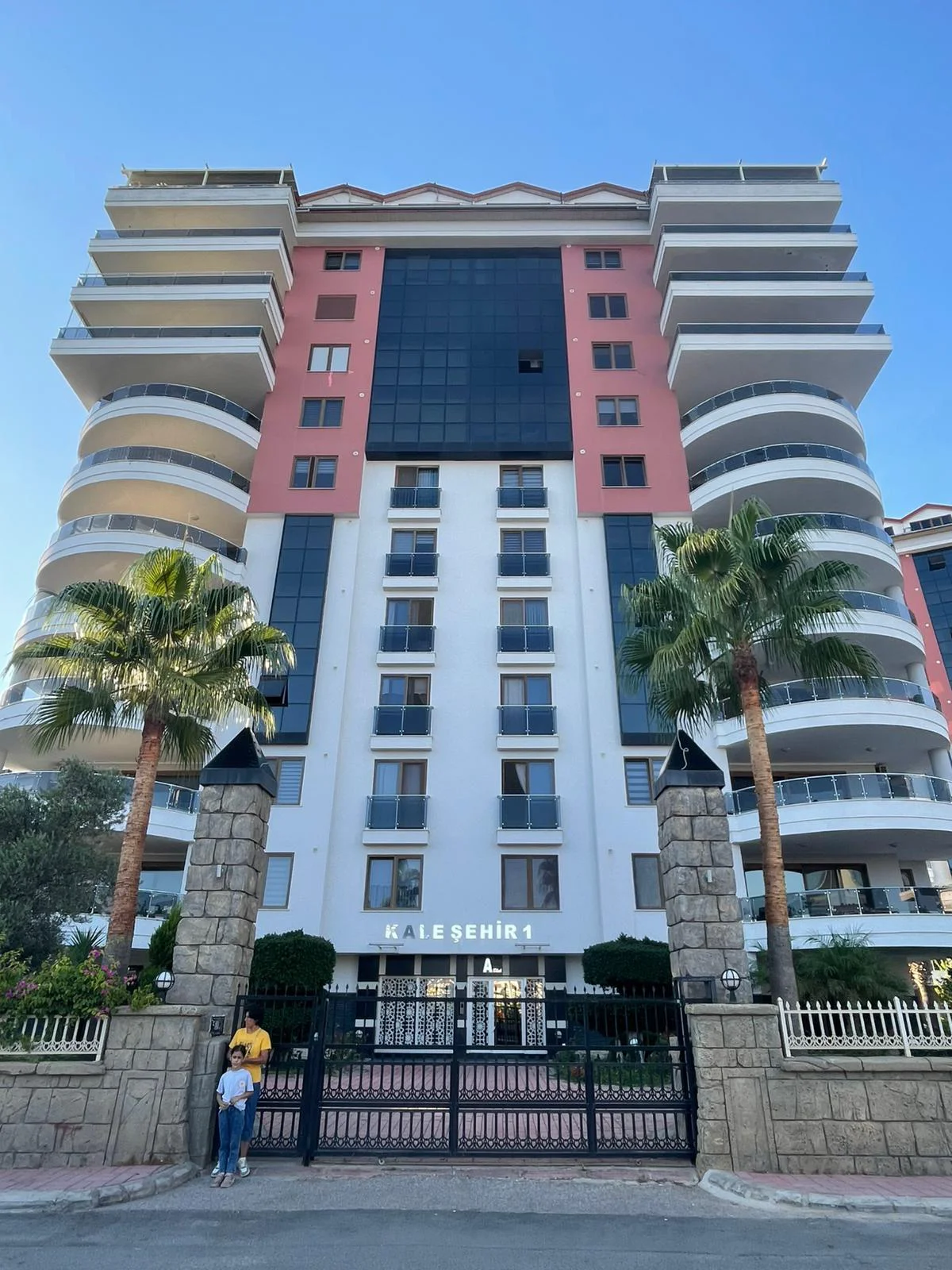 Moderne Außenansicht eines Hochhaus-Apartmentgebäudes mit Balkonen. Außenansicht eines modernen mehrstöckigen Wohngebäudes in der Türkei mit geschwungenen Balkonen, großen Glasfronten und einem gesicherten Eingangsbereich mit Palmen.