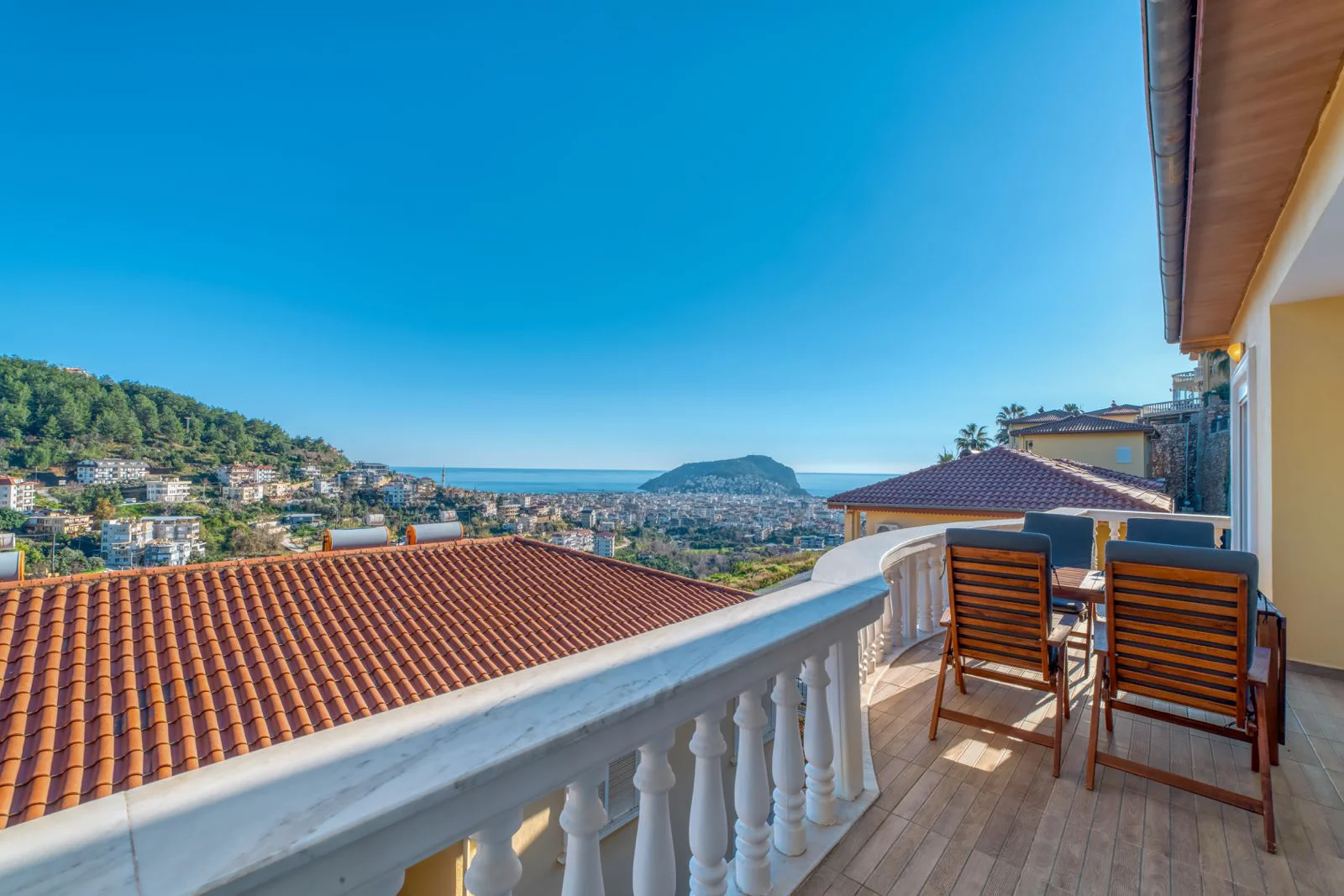 Großzügiger Balkon mit Panoramablick auf die Stadt und das Mittelmeer. Ein heller, geräumiger Balkon mit Bodenbelag in Holzoptik und einer weißen Balustrade. Ausgestattet mit einer Essgruppe im Freien und einem Panoramablick auf die Stadt, das Mittelmeer und grüne Hügel unter einem klaren blauen Himmel.