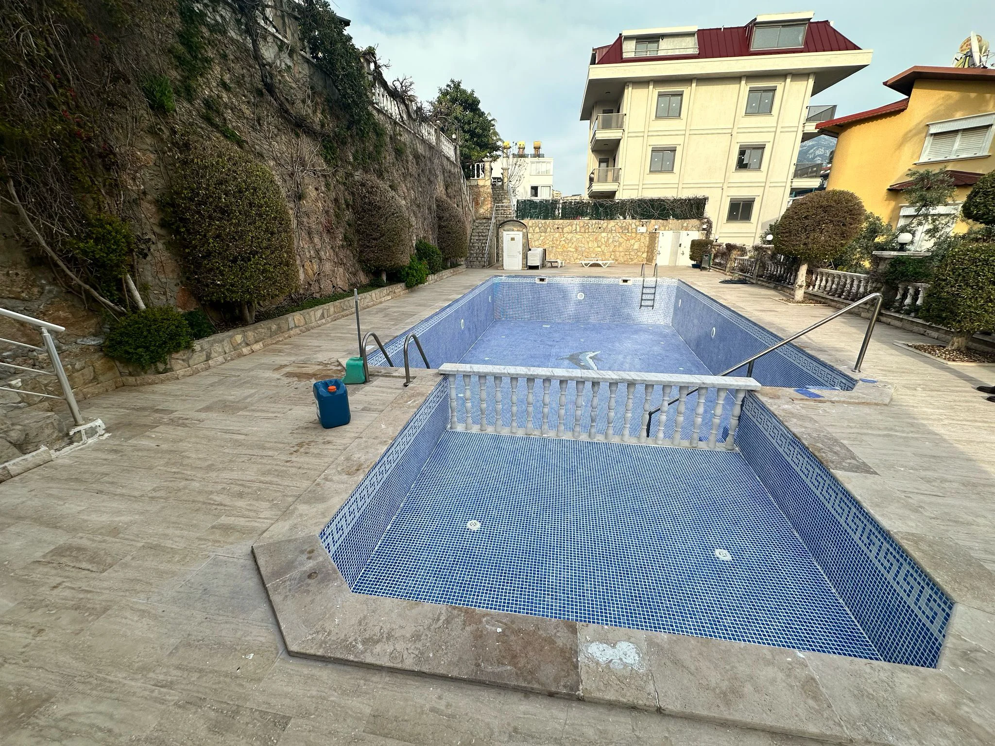 Leerer Außenpool mit Steinterrasse. Gemeinschaftlicher Außenpoolbereich mit zwei leeren Becken mit blauen Mosaikfliesen und einer umgebenden Steinterrasse vor Wohngebäuden. Scheint gewartet zu werden.