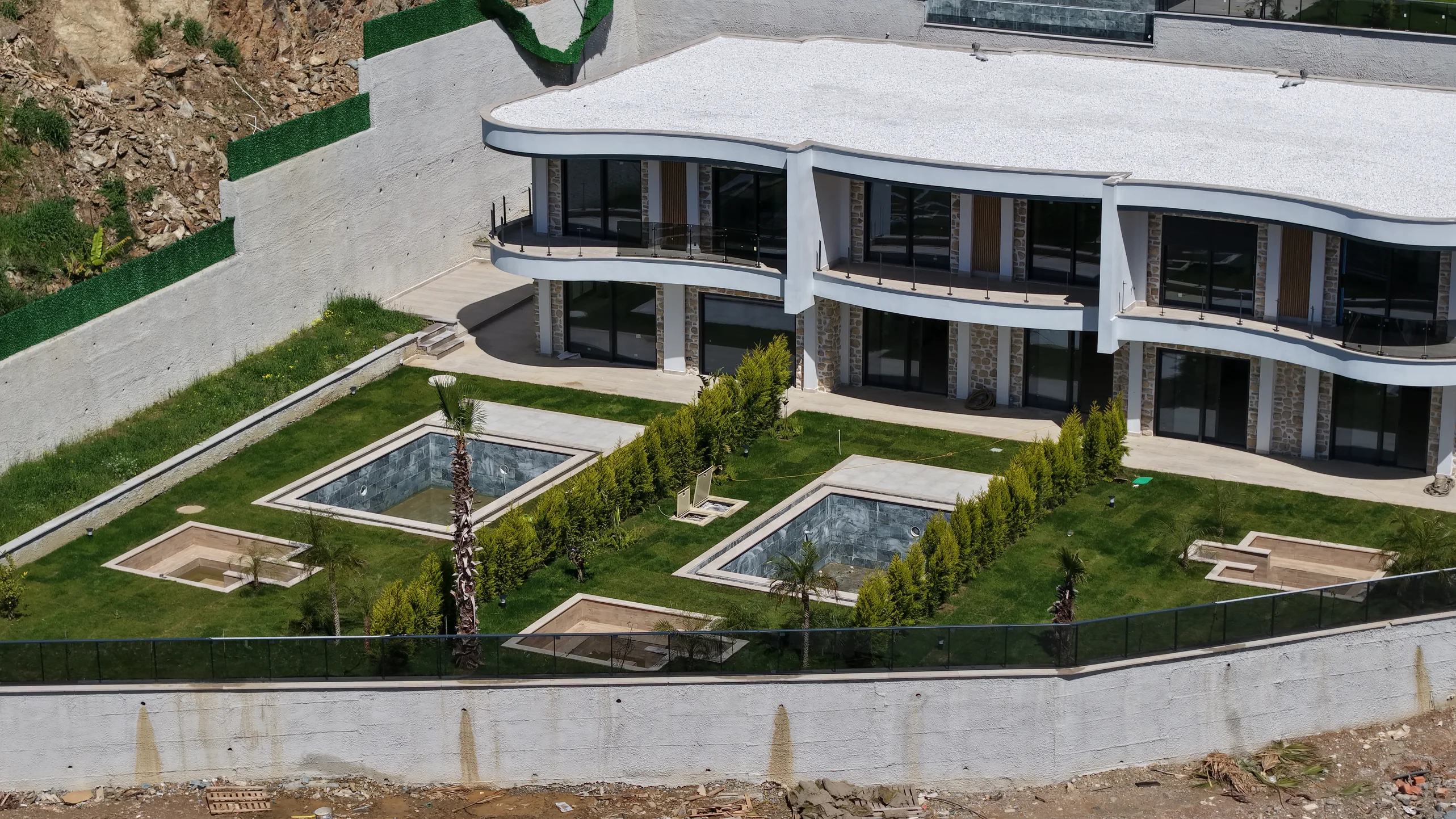 Atemberaubende moderne Villen-Außenansicht mit privatem Garten und Pools. Außenansicht einer modernen Luxusvilla mit wellenförmigem architektonischem Design, Glasbalkonen und einem angelegten grünen Garten mit zwei rechteckigen privaten Pools. Auf einem Hügel gelegen.