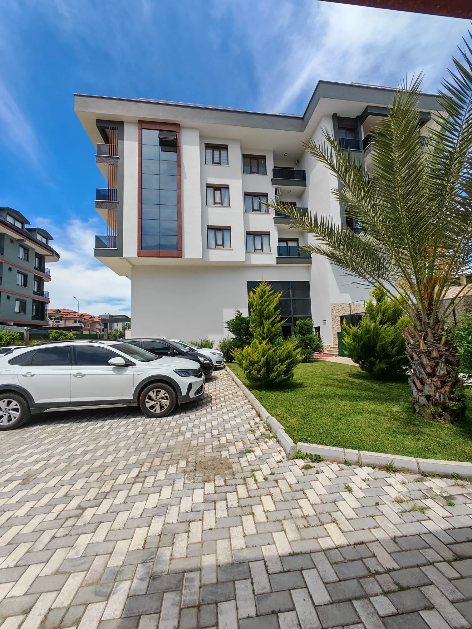 Außenansicht eines modernen Apartmentgebäudes mit Parkplatz und Garten. Außenansicht eines modernen Apartmentgebäudes mit weiß-grauer Fassade, Balkonen und großen Fenstern. Gepflasterter Parkplatz mit Autos und ein kleiner Landschaftsgarten mit Palmen und Sträuchern.