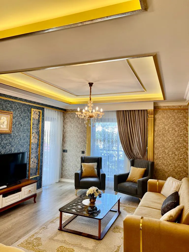 Luxuriöses Wohnzimmer mit Kronleuchter und goldenen Akzenten. Ein luxuriös eingerichtetes Wohnzimmer mit einem goldenen Sofa, zwei Sesseln, einem Glastisch und einem großen Fernseher. Es hat gemusterte Tapeten, Holzböden, einen Kronleuchter und elegante Deckenbeleuchtung.