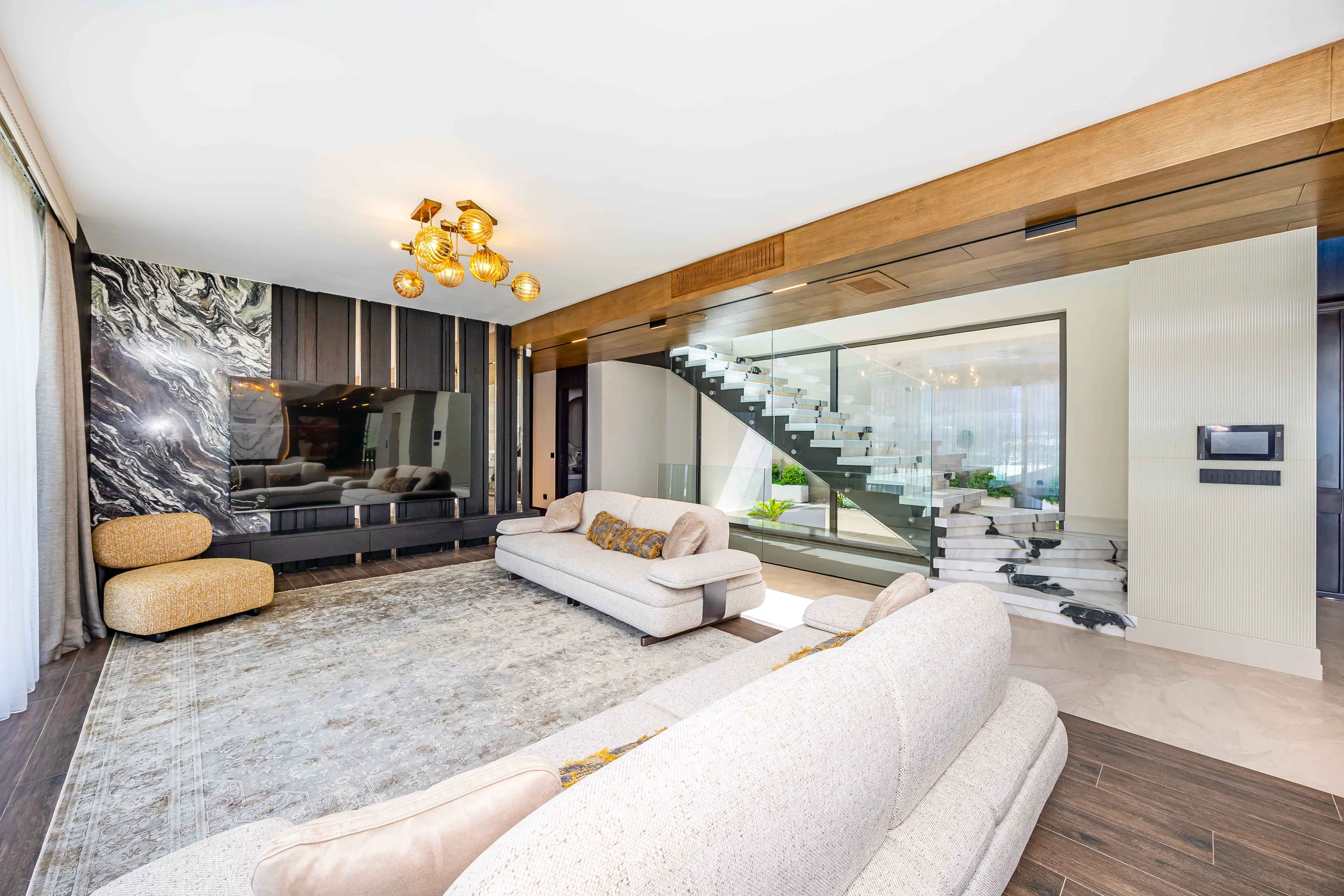 Luxuriöses modernes Wohnzimmer mit Glastreppe. Ein geräumiges, modernes Wohnzimmer mit weißen Sofas, einem Fernseher an einer schwarzen Marmorwand und einer markanten Glastreppe. Mit dunklen Holzböden und einem prunkvollen Kronleuchter.