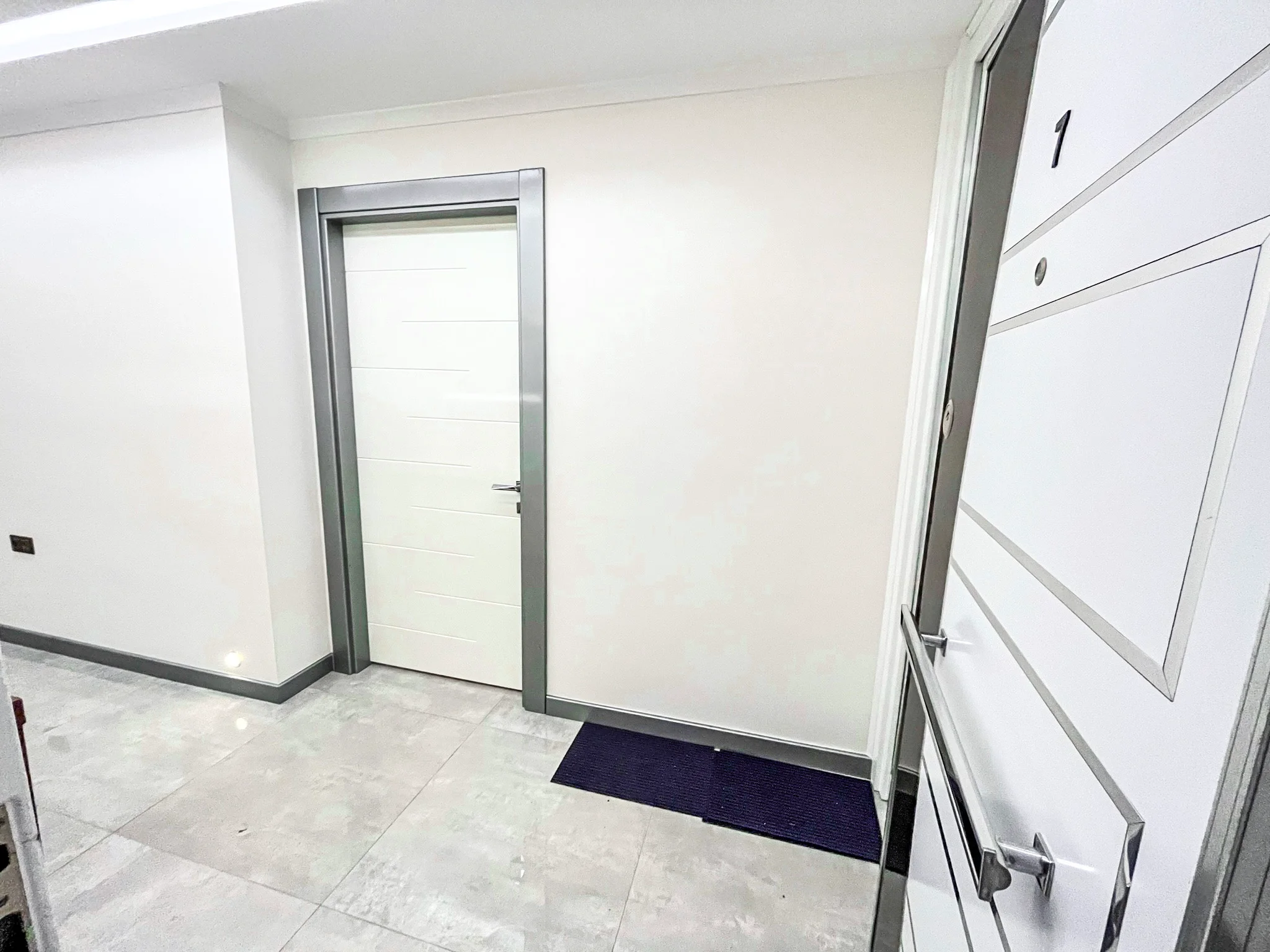 Moderner Wohnungseingangsflur mit Fliesenboden. Ein sauberer und moderner Wohnungseingangsflur mit hellgrauem Fliesenboden und weißen Wänden. Zwei zeitgenössische Türen mit silbernen Akzenten sind sichtbar.