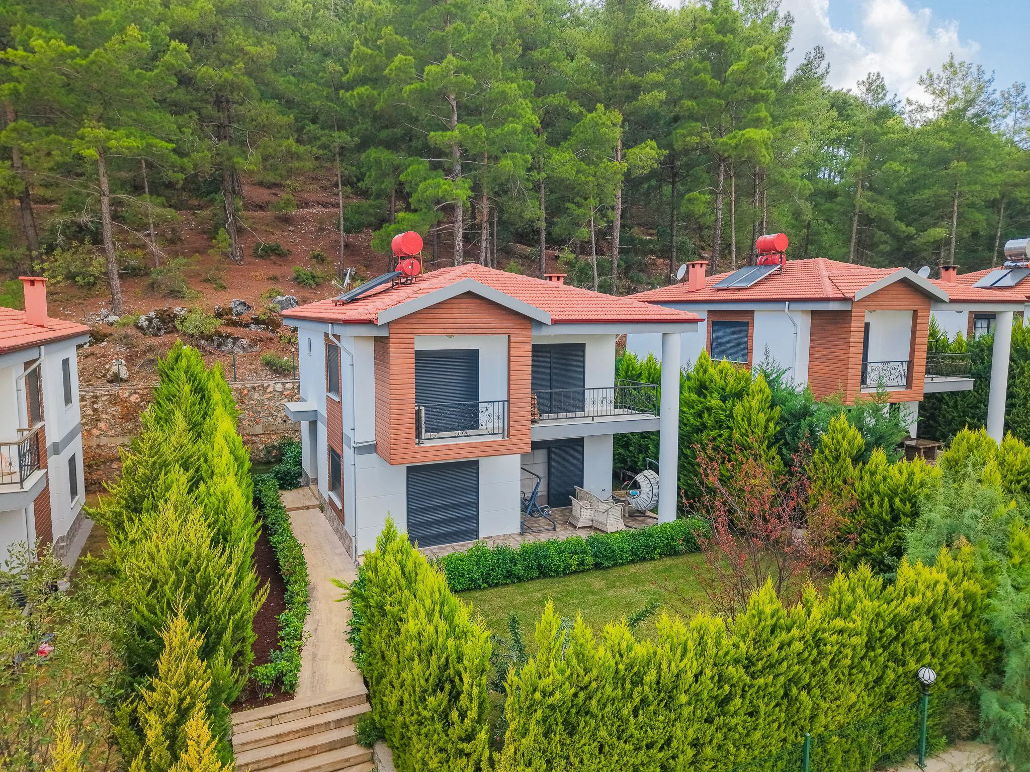 Moderne Villen mit Waldblick und privaten Gärten. Außenansicht moderner, zweistöckiger Villen mit weiß-hölzernen Fassaden, roten Dächern, privaten Gärten und Balkonen. Diese gepflegten Immobilien bieten einen herrlichen Waldblick und Sitzgelegenheiten im Freien.
