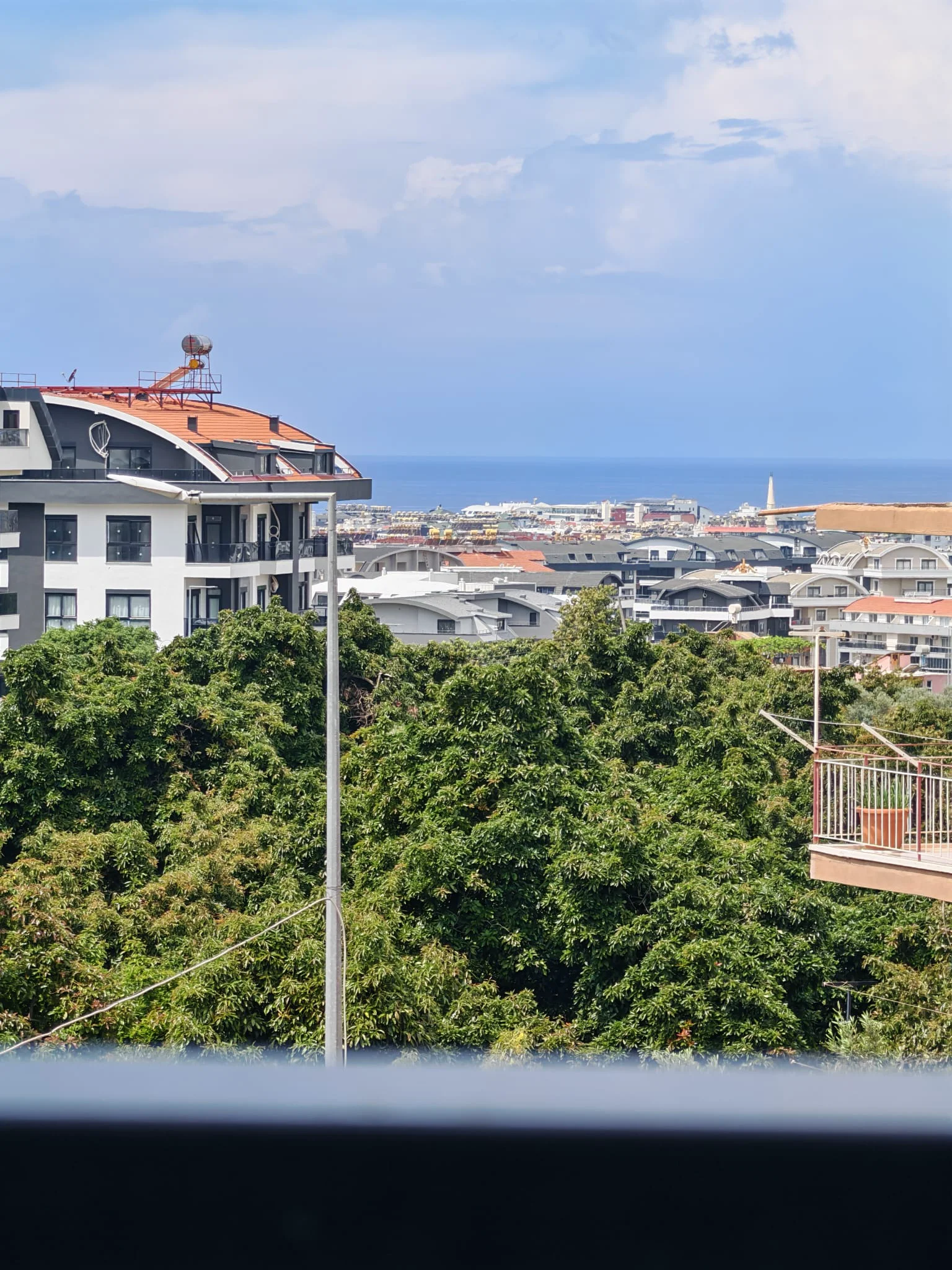 Panoramablick auf Stadt und Mittelmeer. Dieses Bild zeigt eine malerische Aussicht von einem Balkon auf üppige grüne Bäume, eine moderne Stadt und das ferne blaue Mittelmeer unter einem bewölkten Himmel.