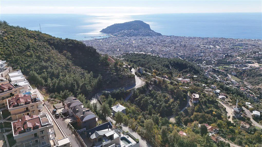 Panoramablick auf Meer und Stadt von den Alanya Mountain Homes. Luftaufnahme moderner Luxusimmobilien an einem grünen Berghang mit kurvigen Straßen, privaten Pools und einem beeindruckenden Panoramablick auf die Stadt Alanya und das glitzernde Mittelmeer.