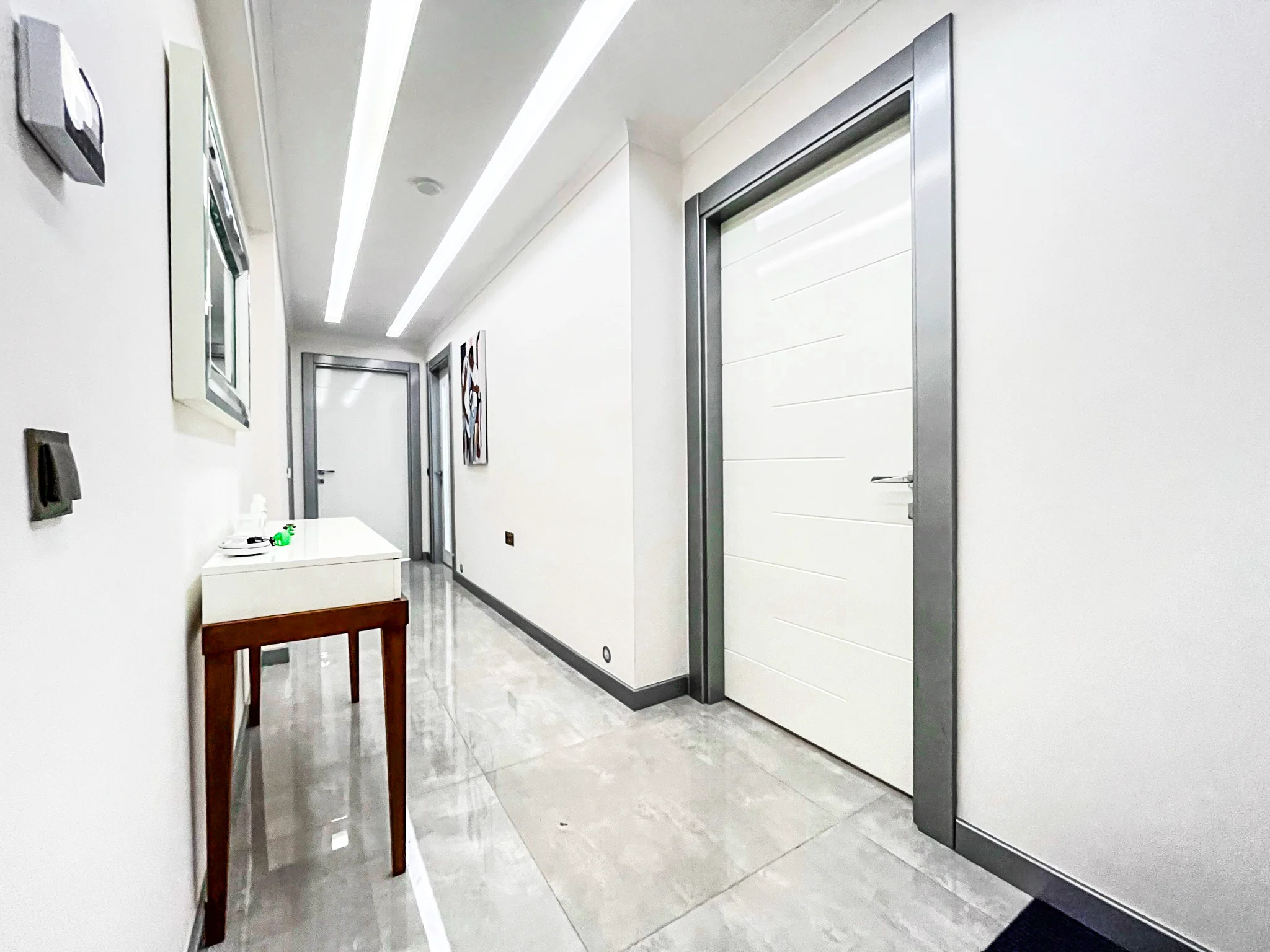Heller moderner Flur mit stilvoller Beleuchtung und Fliesenboden. Ein gepflegter, moderner Flur mit eleganten grauen Bodenfliesen, hellen LED-Lichtleisten und weißen Wänden. Ein kleiner Konsolentisch mit Spiegel setzt einen stilvollen Akzent und führt zu mehreren Türen.