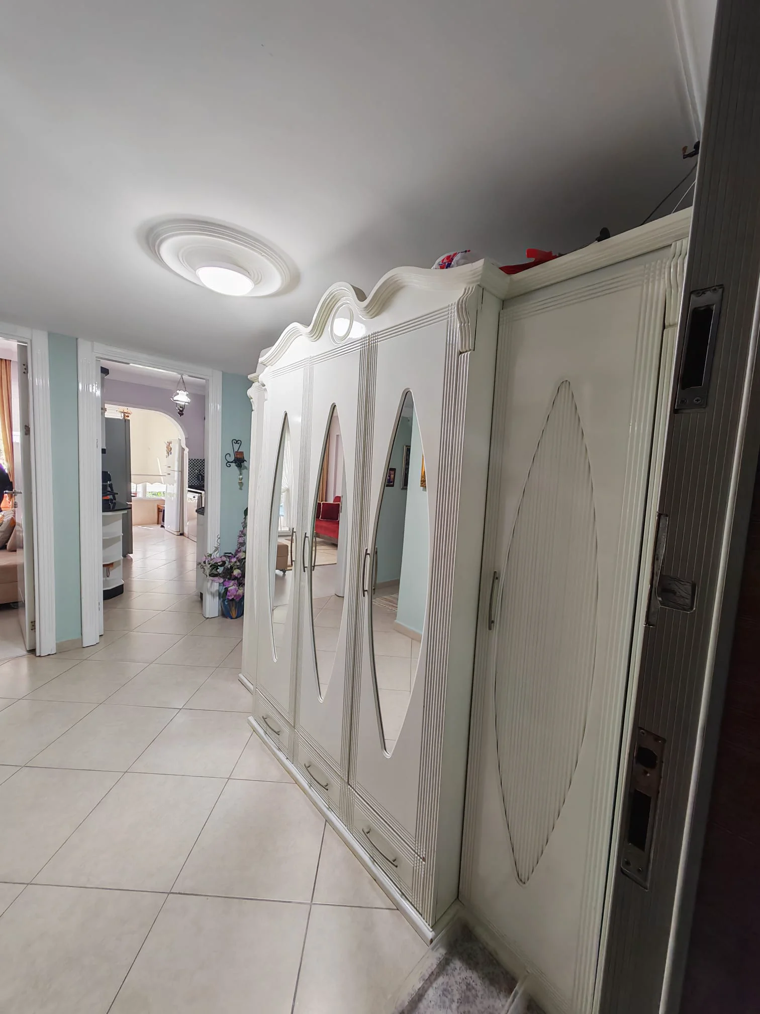 Eleganter weißer verzierter Kleiderschrank im Innenflur. Ein traditioneller Flur, der einen großen, verzierten weißen Kleiderschrank mit Spiegeltüren und Schubladen zeigt. Der Raum verfügt über hell geflieste Böden und eine Deckenleuchte und führt zu weiteren Räumen.