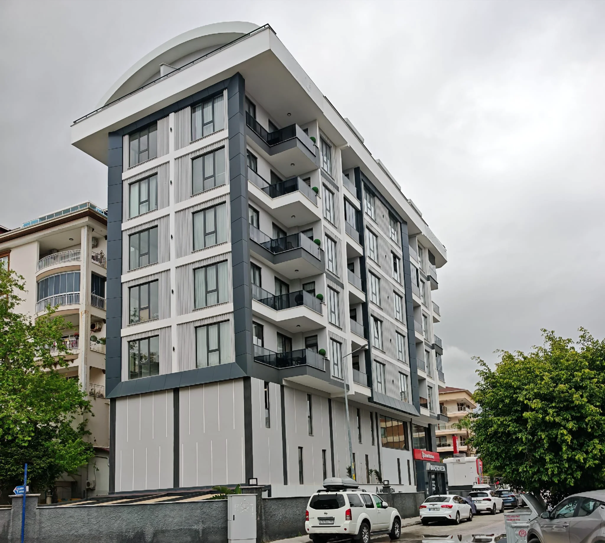 Moderne Wohnhausfassade mit Balkonen in städtischer Umgebung. Außenansicht eines modernen mehrstöckigen Wohngebäudes in zeitgenössischem Design mit grau-weißen Fassaden, Balkonen mit Glasgeländern und großen Fenstern. Autos parken an der Straße.