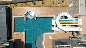 Luftaufnahme eines modernen Swimmingpools mit Wasserrutsche. Luftaufnahme eines modernen Resort-Swimmingpools mit einem großen blauen Becken, einer weißen Wasserrutsche mit Regenbogenstreifen und einem sprudelnden Whirlpool. Dieser lebhafte Wasserbereich ist sehr gepflegt.