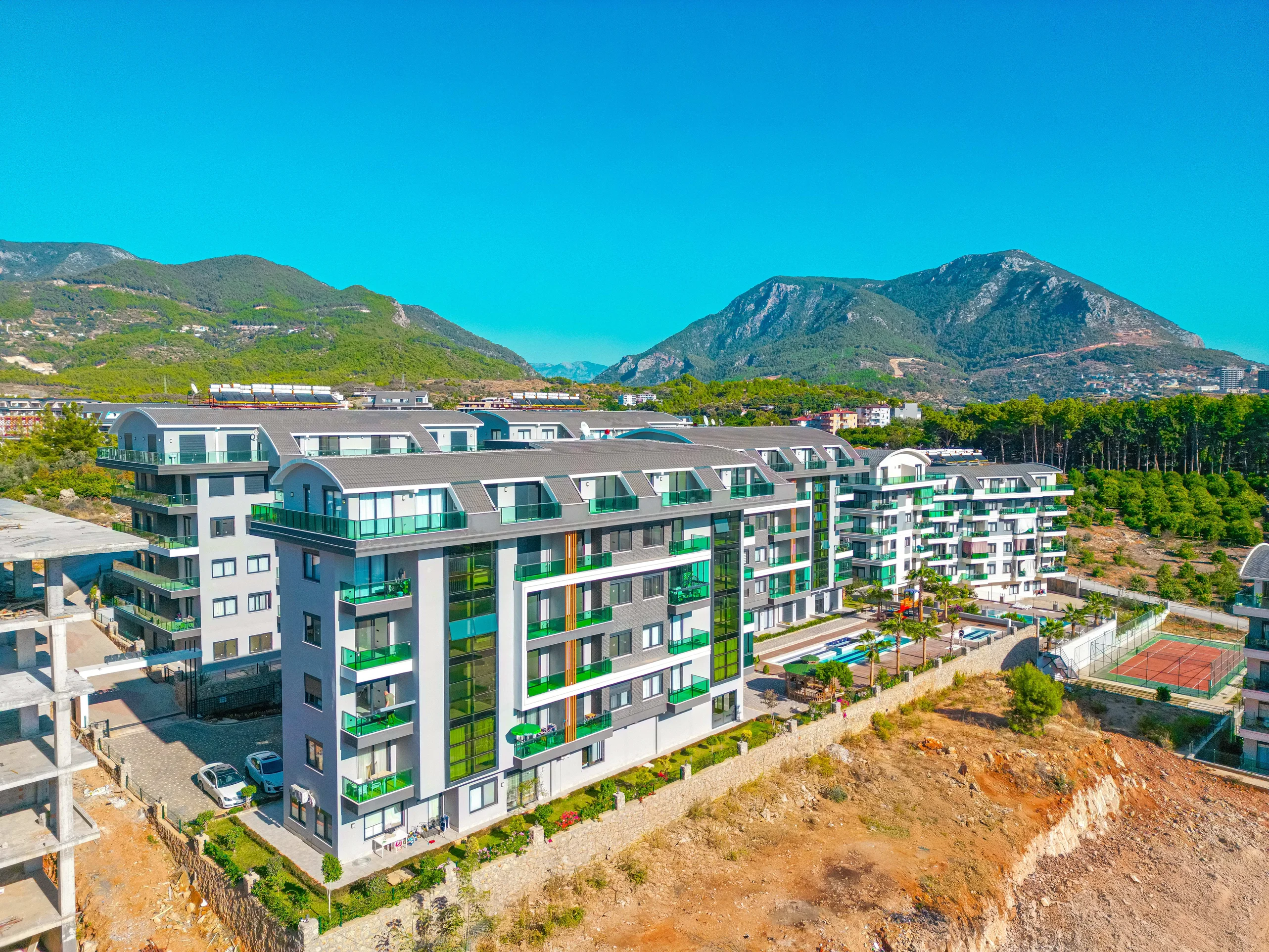 Moderner Apartmentkomplex mit atemberaubendem Bergblick. Außenansicht einer modernen Wohnanlage mit Balkonen mit grünen Geländern, Swimmingpool und Tennisplatz. Majestätische grüne Berge und ein klarer blauer Himmel bilden eine malerische Kulisse.