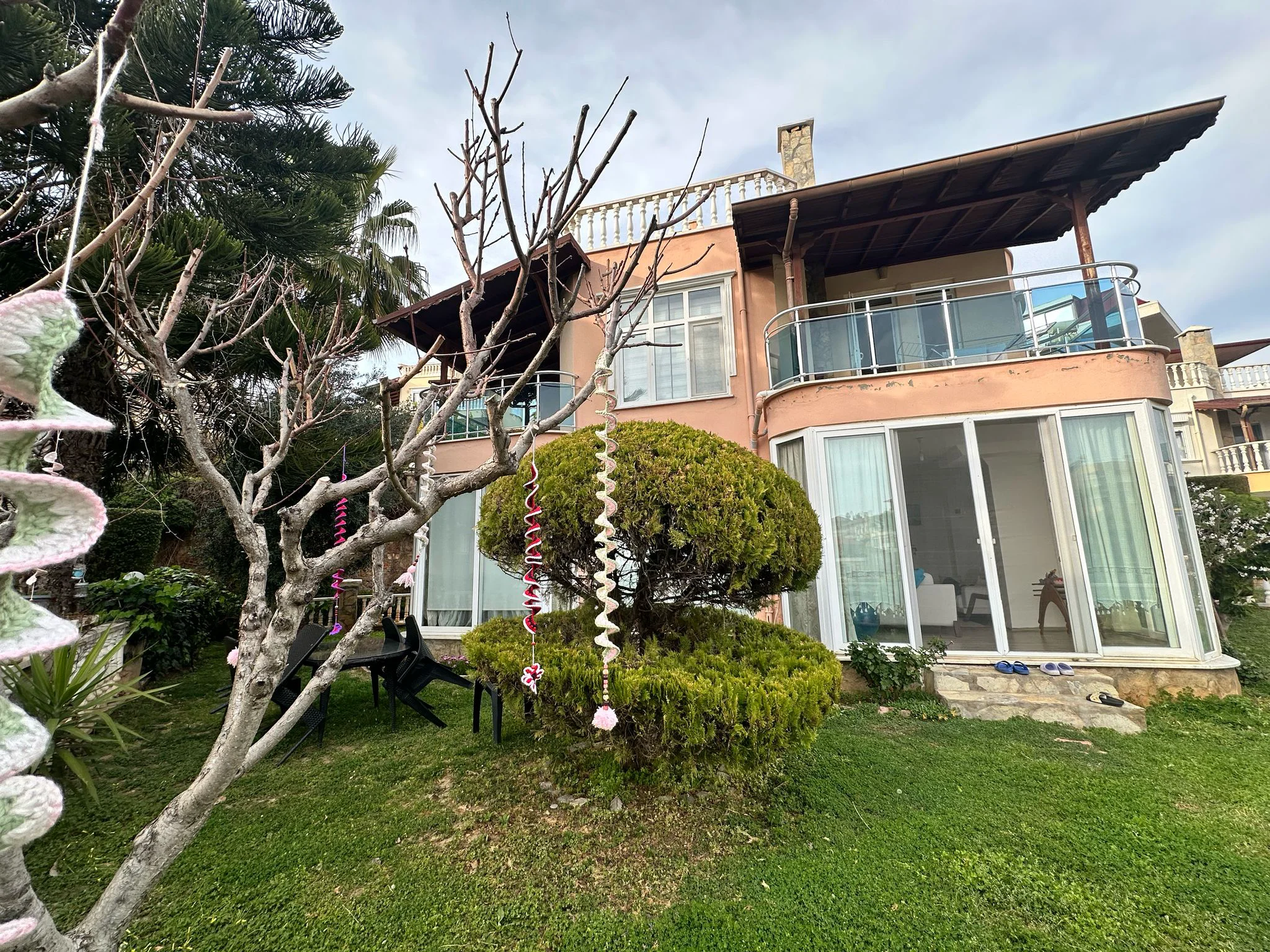 Charmante Außenansicht eines Wohnhauses mit Garten und Balkon. Außenansicht eines mehrstöckigen Wohnhauses mit rosa Fassade, einem üppigen grünen Garten und einem modernen Balkon mit Glasgeländer und Holzpergola. Zeigt den Außenwohnbereich.