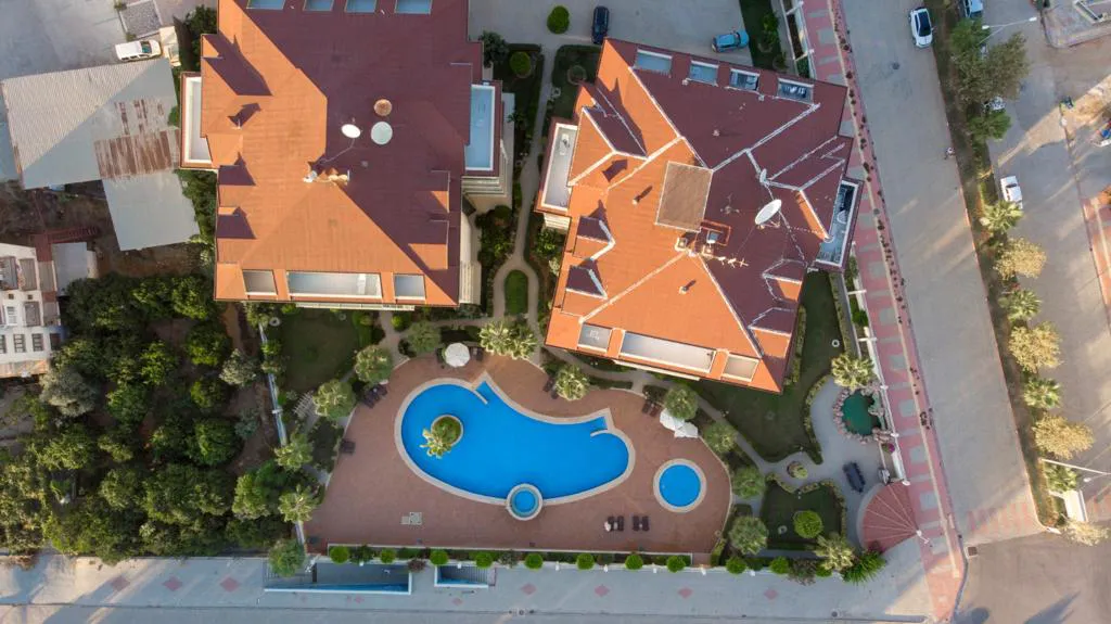 Luxuriöse Wohnanlage mit großem Außenpool. Eine Luftaufnahme einer modernen Wohnanlage. Sie zeigt zwei große Gebäude mit roten Dächern, einen großen, unregelmäßig geformten Swimmingpool, grüne Landschaftsgärten und Sonnenliegen für Bewohner.