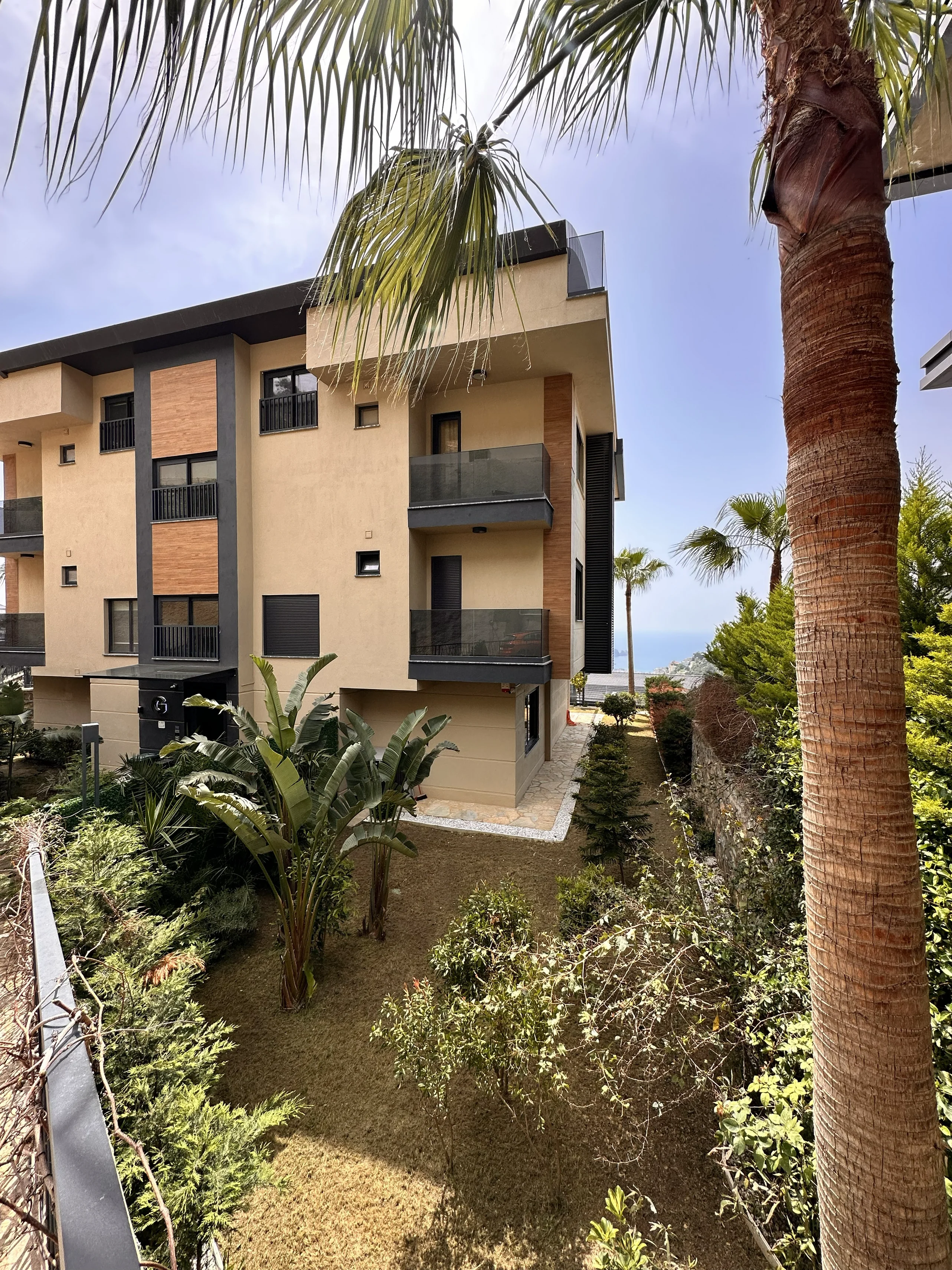 Modernes Apartmentgebäude mit üppigem Garten und Meerblick. Außenansicht eines modernen Apartmentgebäudes mit mehreren Balkonen mit Glasgeländer und einem großen, gepflegten Garten mit Palmen. Ein ferner Meerblick ist unter klarem Himmel sichtbar.