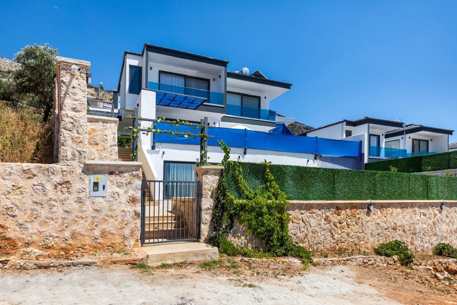Moderne Hangvilla-Außenansicht mit blauen Balkonen. Außenansicht einer modernen mehrstöckigen Villa am Hang mit weißen Wänden, blauen Sichtschutzwänden und Glasbalkonen. Mit rustikalem Steinzaun, Metalltor und grünen künstlichen Hecken, die zeitgenössisches Design präsentieren.