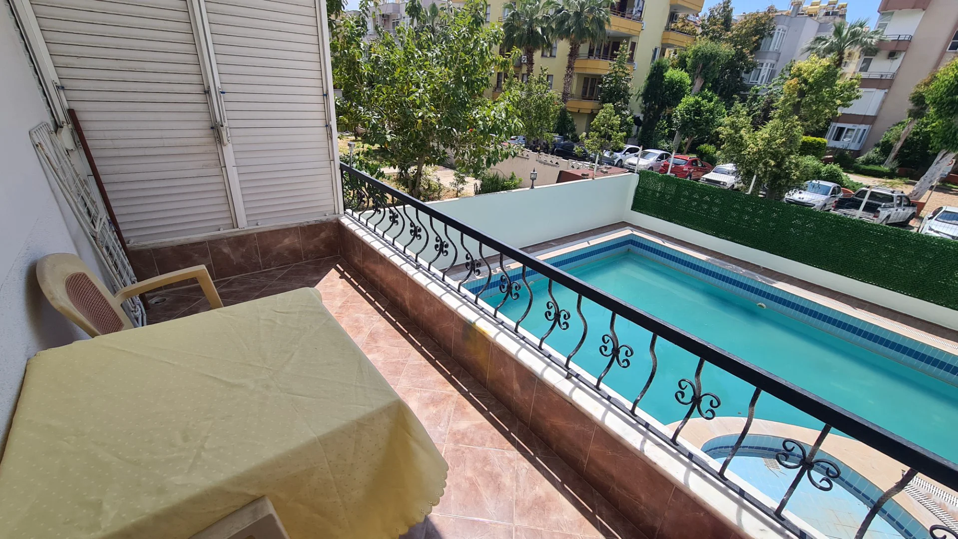 Sonniger Balkon mit Poolblick und Sitzgelegenheiten im Freien. Ein sonniger Balkon mit Terrakottafliesen und einem verzierten Metallgeländer. Er verfügt über einen Außentisch und einen Stuhl und bietet einen angenehmen Blick auf den Swimmingpool der Anlage und den umliegenden grünen Garten.
