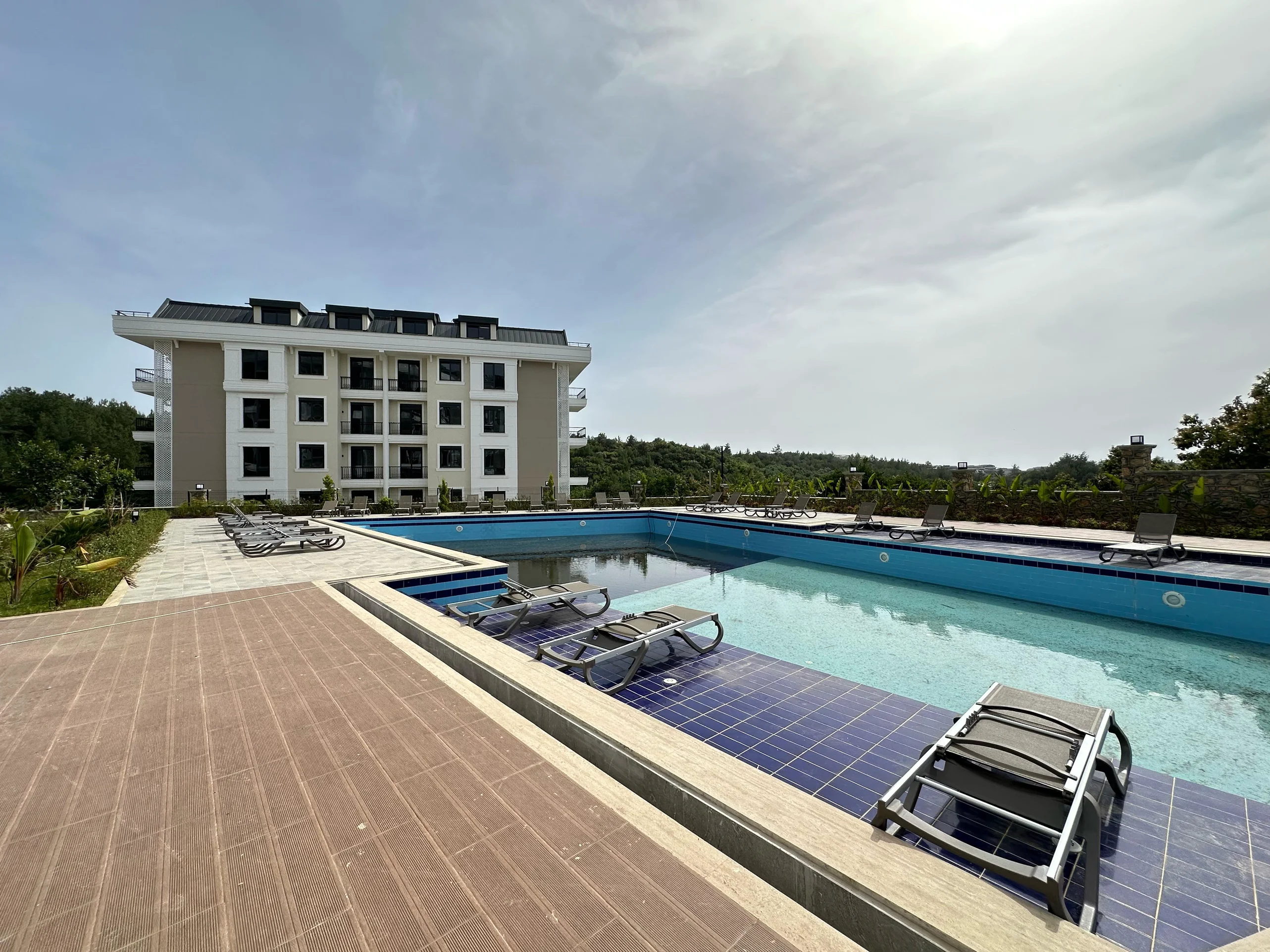Modernes Apartmenthaus mit großem Außenpool. Außenpoolbereich einer modernen Wohnanlage. Er verfügt über einen großen Pool mit blauen Fliesen, mehrere Sonnenliegen und ein modernes Apartmentgebäude, umgeben von grüner Landschaftsgestaltung.