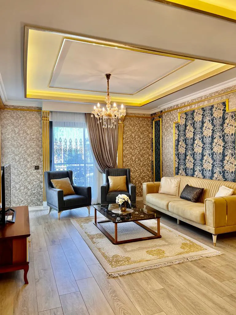 Geräumiges modernes Wohnzimmer mit stilvollen Goldakzenten. Ein modernes und elegantes Wohnzimmer mit hellem Holzboden, goldakzentuierten Wänden, einem großen Kronleuchter und bequemen Sitzgelegenheiten mit Blick ins Grüne.