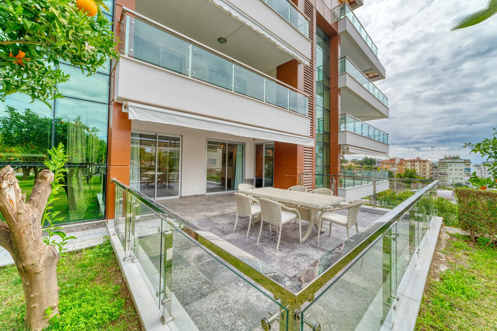 Moderne Erdgeschoss-Terrasse mit Essbereich im Freien. Moderne Erdgeschoss-Terrasse mit gefliestem Innenhof und Essbereich im Freien, umgeben von einem grünen Rasen und Glasgeländern. Verfügt über ein Vordach und Blick auf die Stadt.