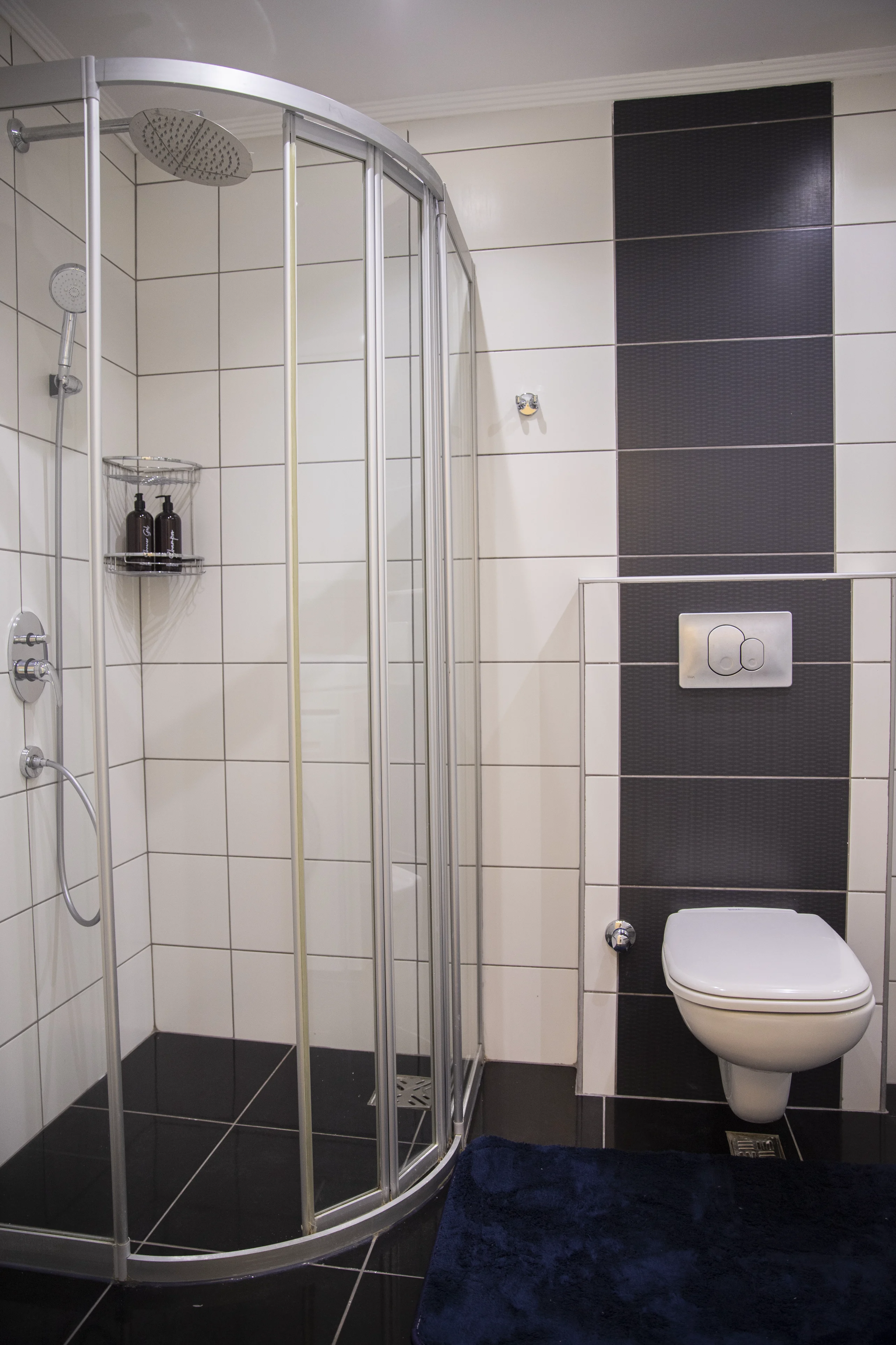 Modernes Badezimmer mit eleganter Duschkabine und Wand-WC. Ein modernes, gepflegtes Badezimmer mit einer gebogenen Glasduschkabine, einem Regenduschkopf und einem Wand-WC. Es verfügt über stilvolle schwarz-weiß geflieste Wände und Böden.