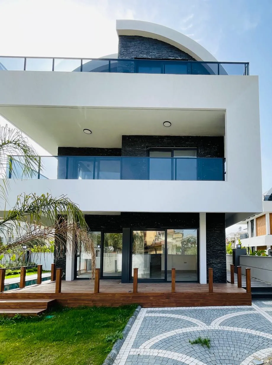 Moderne Luxusvilla-Außenansicht mit Balkonen und Terrasse. Moderne Villa-Außenansicht mit weißer und dunkler Steinfassade, Glasbalkonen und großen Schiebetüren. Mit Holzterrasse, grünem Rasen und gemustertem Kopfsteinpflasterweg.
