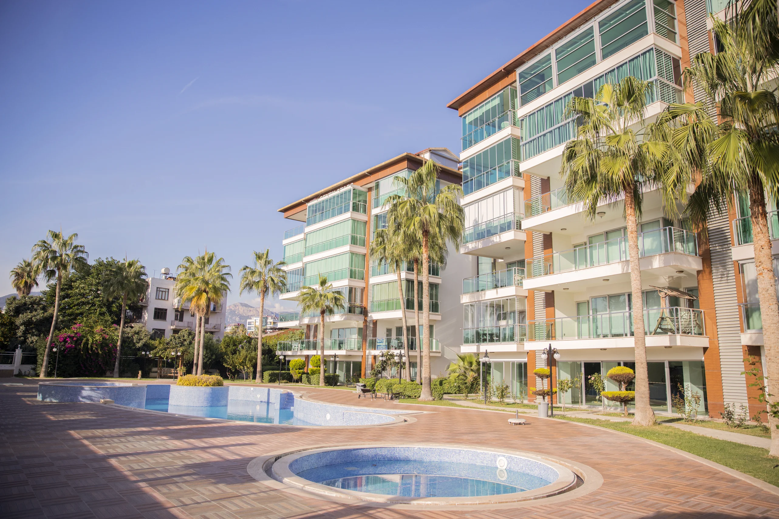 Moderner Wohnkomplex mit Außenpool und Palmen. Außenansicht eines modernen Wohnkomplexes mit großen und runden Schwimmbecken, umgeben von Palmen. Zeitgenössische Gebäude mit Glasbalkonen sind unter einem klaren blauen Himmel gut gepflegt.
