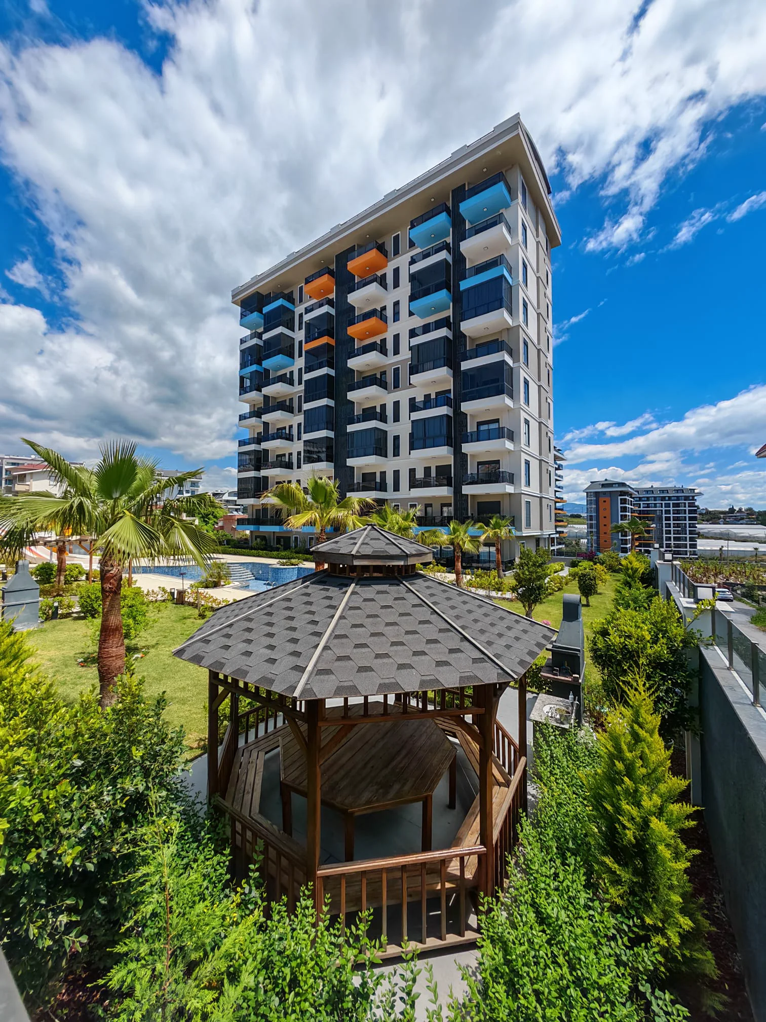 Modernes Gebäude mit Garten, Pavillon und Pool. Außenansicht eines modernen Apartmentkomplexes mit einem Holzpavillon, einem üppig grünen Garten mit Palmen und einem Swimmingpool, ideal für soziale Aktivitäten im Freien und Entspannung.