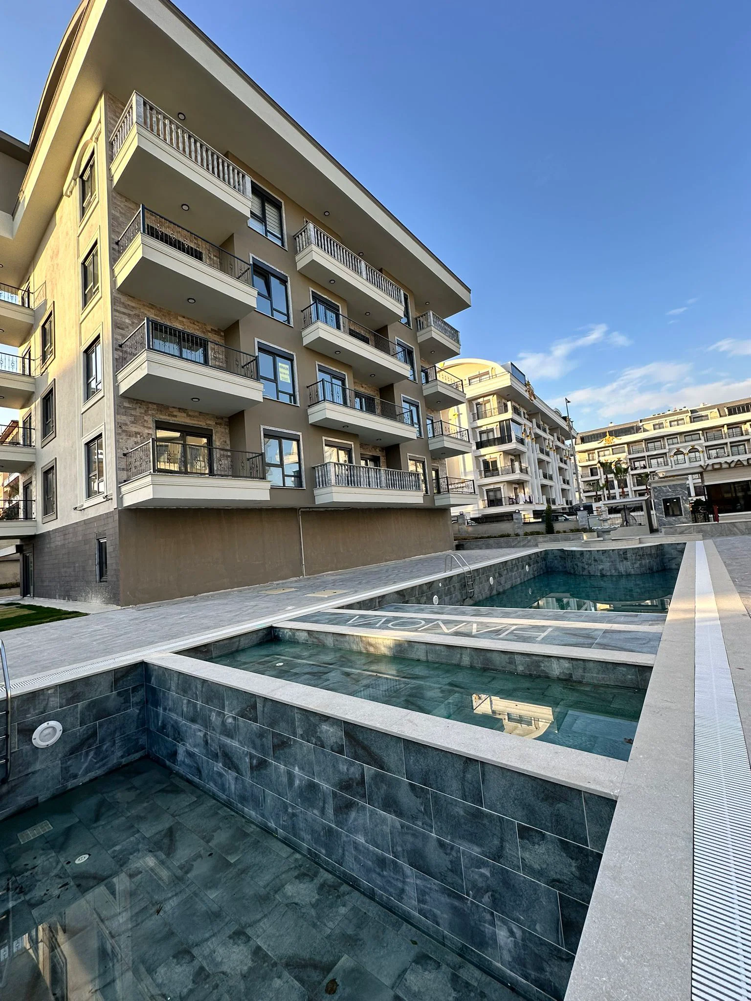 Moderner Apartmentkomplex mit Außenpools und Balkonen. Außenansicht eines modernen Apartmentkomplexes mit zeitgenössischer Architektur, zahlreichen privaten Balkonen und mehrstöckigen Außenpools mit gefliesten Umrandungen unter klarem Himmel.