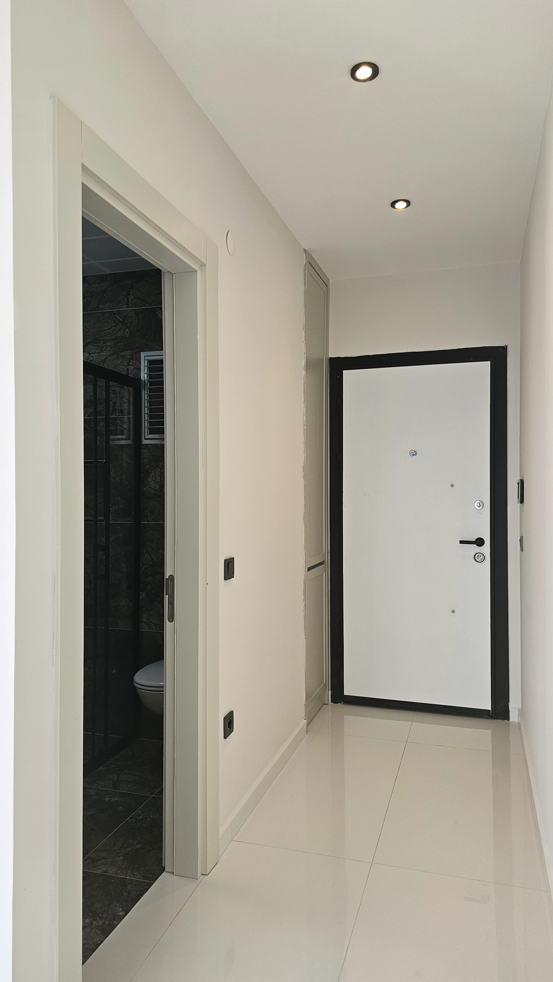 Moderner Eingangsbereich mit Einbauleuchten. Moderner Flur mit glänzend weißem Fliesenboden, weißen Wänden und Einbauleuchten. Verfügt über eine weiße Eingangstür und einen Blick in ein angrenzendes Badezimmer mit dunklen Fliesen.