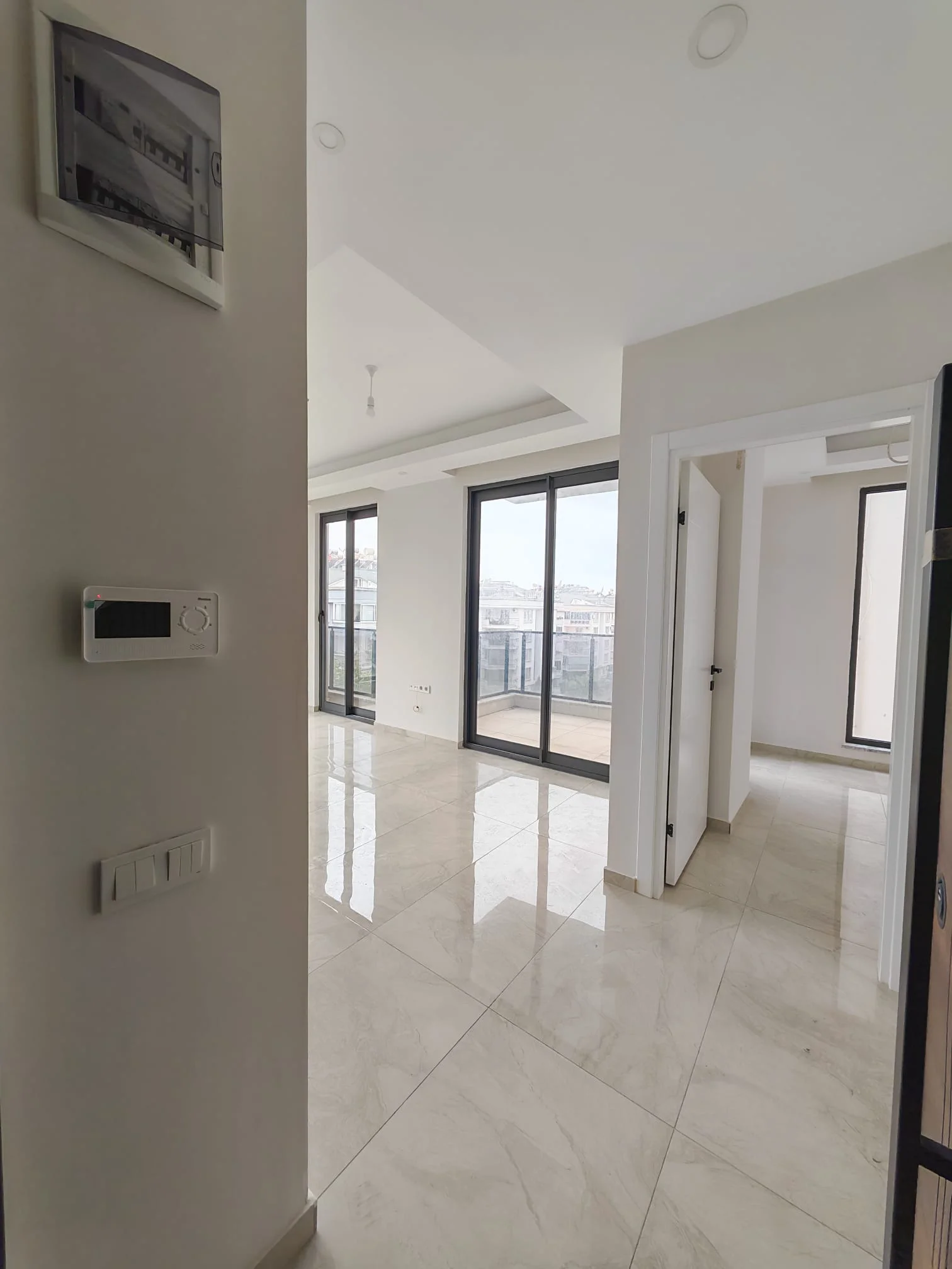 Helles modernes Wohnzimmer mit Balkonzugang. Ein helles, modernes Wohnzimmer mit glänzenden, hellen Fliesenböden und weißen Wänden. Große Glasschiebetüren führen zu einem Balkon mit Stadtblick. Der Raum wirkt neu renoviert und sauber, mit Einbaustrahlern.