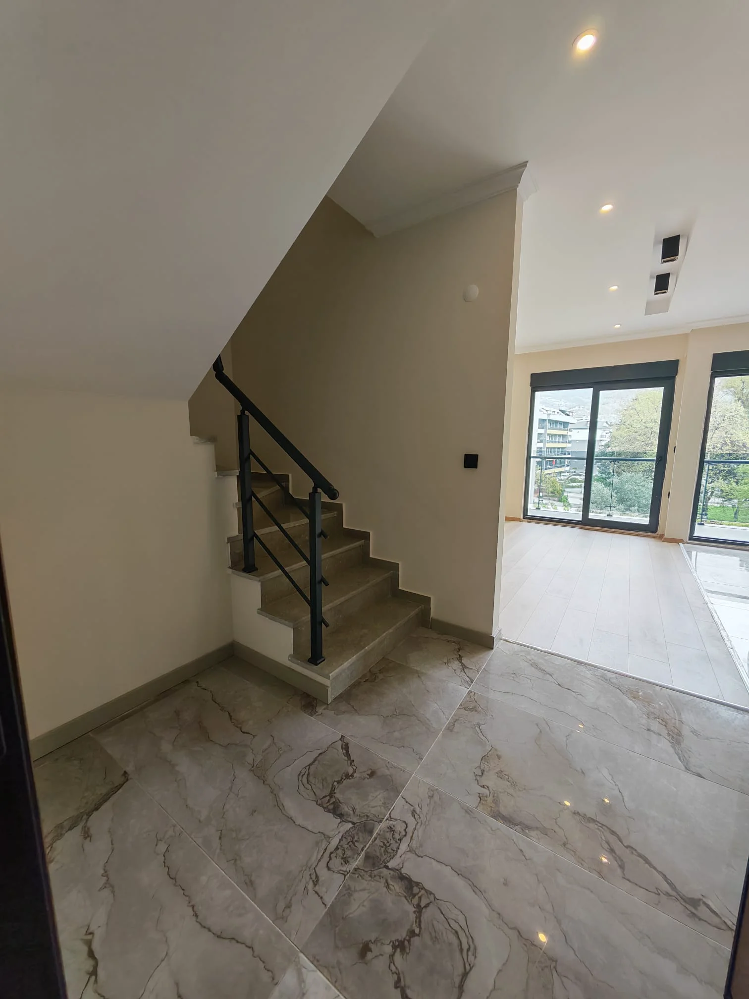 Moderner Eingangsbereich mit eleganter Treppe und hellem Wohnbereich. Heller, moderner Eingangsbereich mit großen Bodenfliesen in Marmoroptik und einer stilvollen Treppe mit schwarzem Metallgeländer. Führt zu einem geräumigen Wohnbereich mit Zugang zum Balkon.