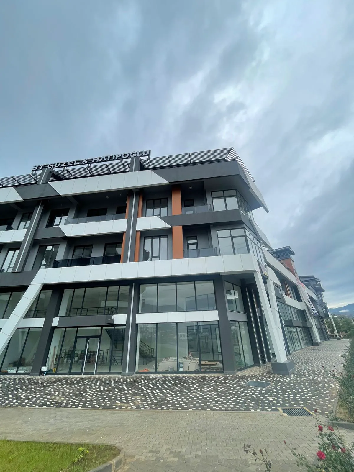 Neues modernes gemischt genutztes Gebäude mit elegantem architektonischem Design. Außenansicht eines modernen, mehrstöckigen Gebäudes mit gemischter Nutzung, das sich durch elegante, winklige Architektur, große Schaufenster, Balkone in den oberen Etagen und einen gepflasterten Eingang unter bewölktem Himmel auszeichnet.