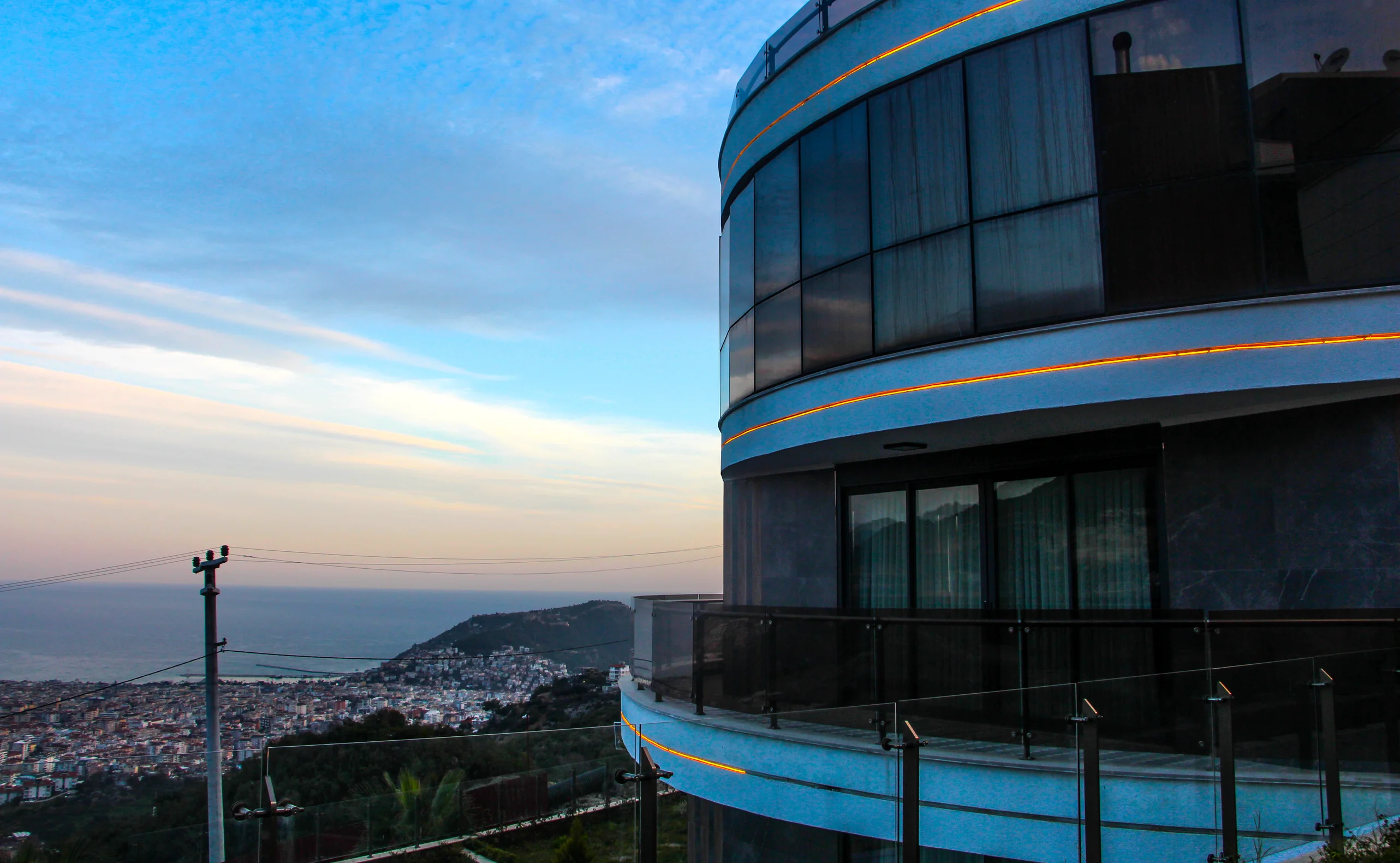 Moderne Gebäudeterrasse mit Panorama-Meer- und Stadtblick. Eine moderne, geschwungene Gebäudeterrasse mit eleganten Glasgeländern und beleuchteten Vorsprüngen, die einen atemberaubenden Panoramablick auf das ferne Meer und eine lebendige Stadt darunter bietet.