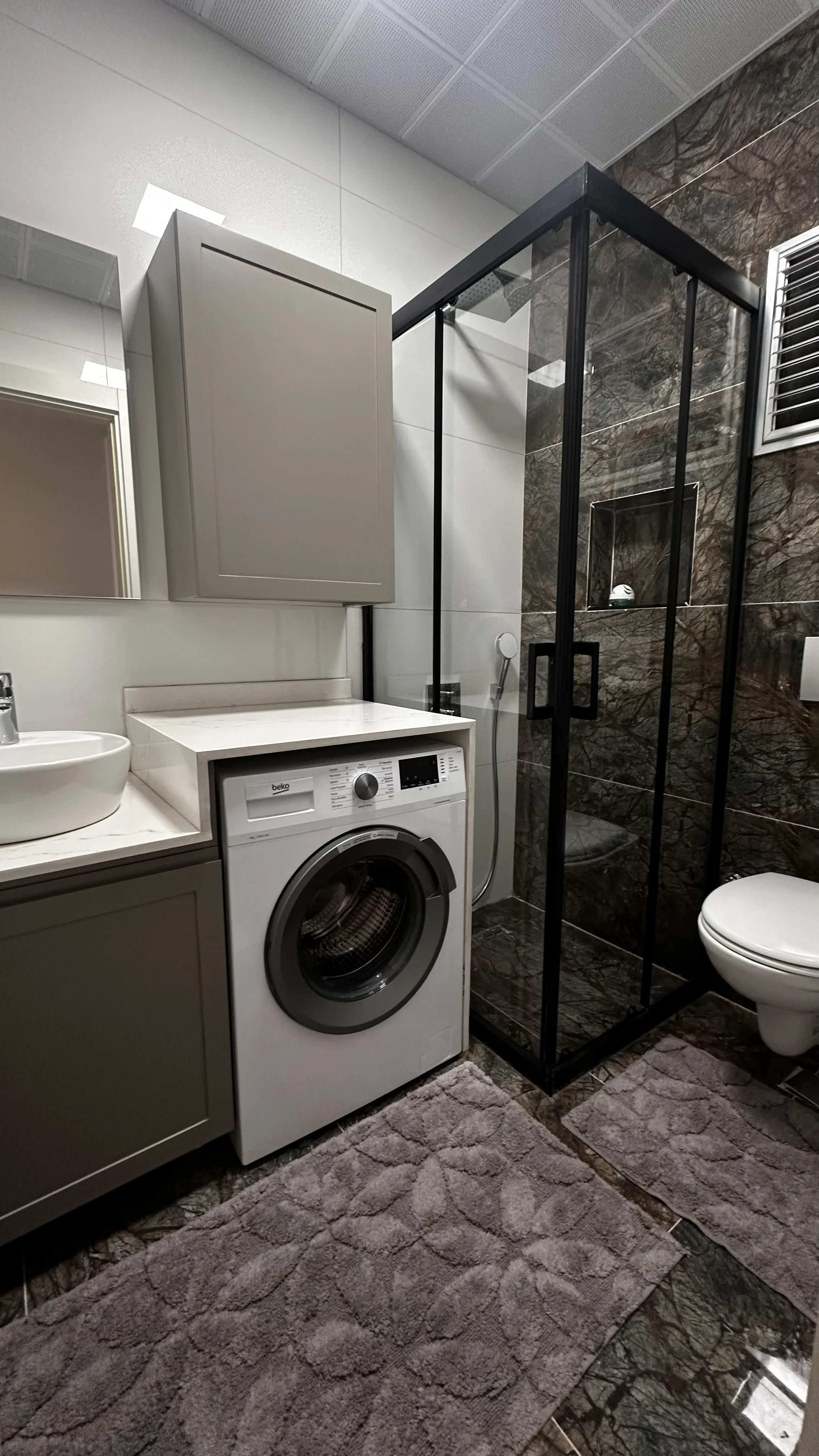 Modernes Badezimmer mit Waschmaschine und Glasdusche. Ein modernes, stilvolles Badezimmer mit dunklen Fliesen in Marmoroptik, einer Glasdusche mit schwarzem Rahmen, einer wandhängenden Toilette, einem Waschtisch mit weißem Aufsatzwaschbecken und einer integrierten Waschmaschine.