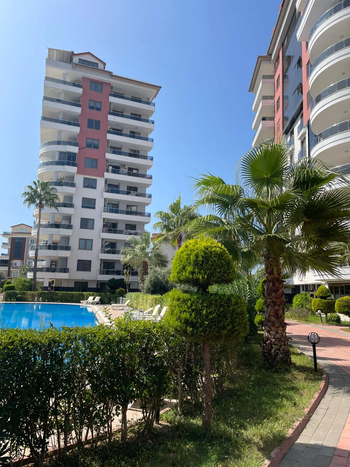Moderner Apartmentkomplex mit großem Außenpool. Außenansicht einer modernen Wohnanlage mit einem großen blauen Swimmingpool, Sonnenliegen und tropischen Palmen unter einem klaren, sonnigen Himmel. Gepflegte Gärten und Wege.