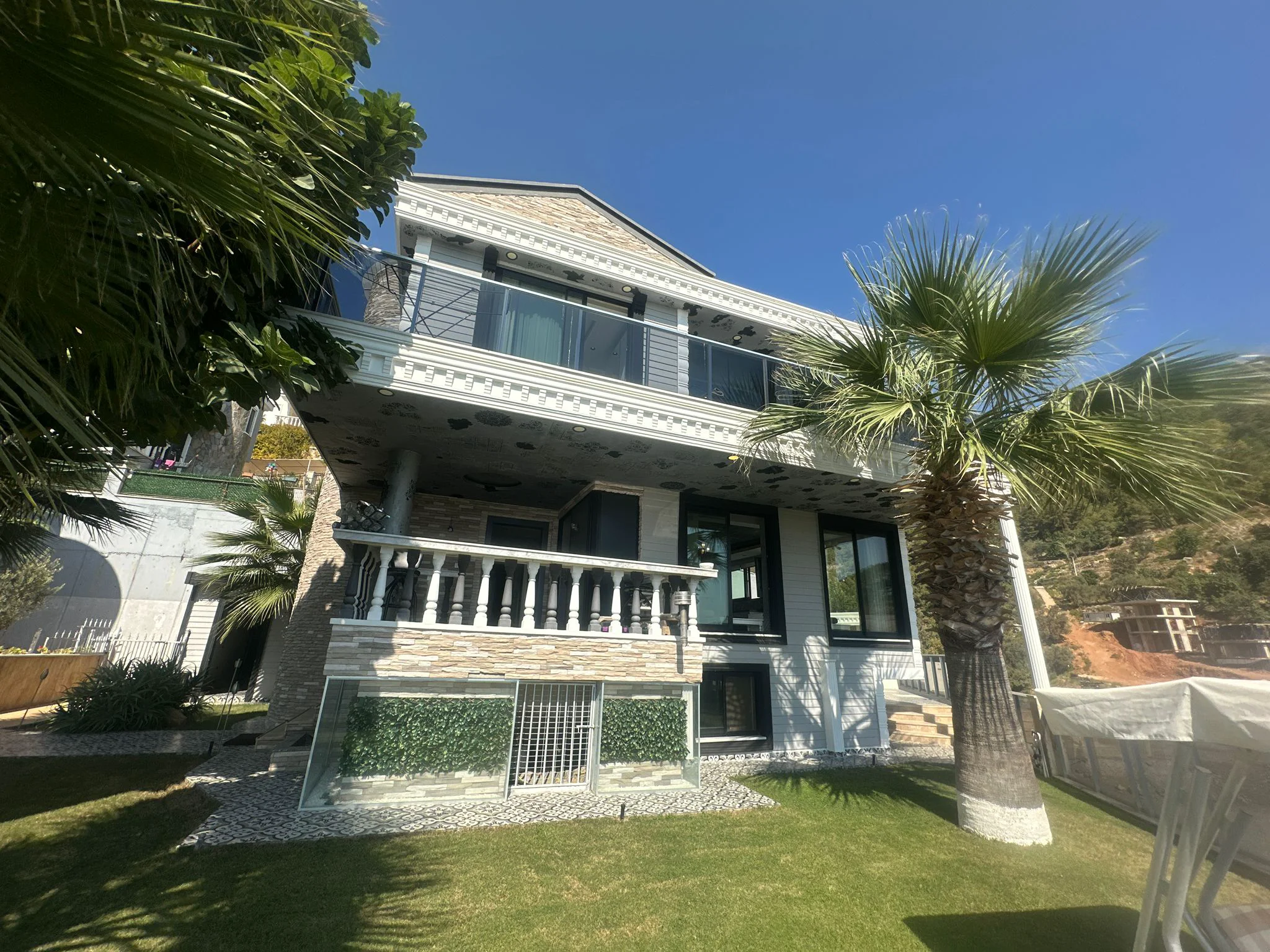 Moderne mehrstöckige Villa Außenansicht mit üppigem Garten und Balkonen. Außenansicht einer modernen mehrstöckigen Villa mit Balkonen, Steinfassade und großen Fenstern. Mit gepflegtem grünem Rasen und Palmen unter klarem blauem Himmel.