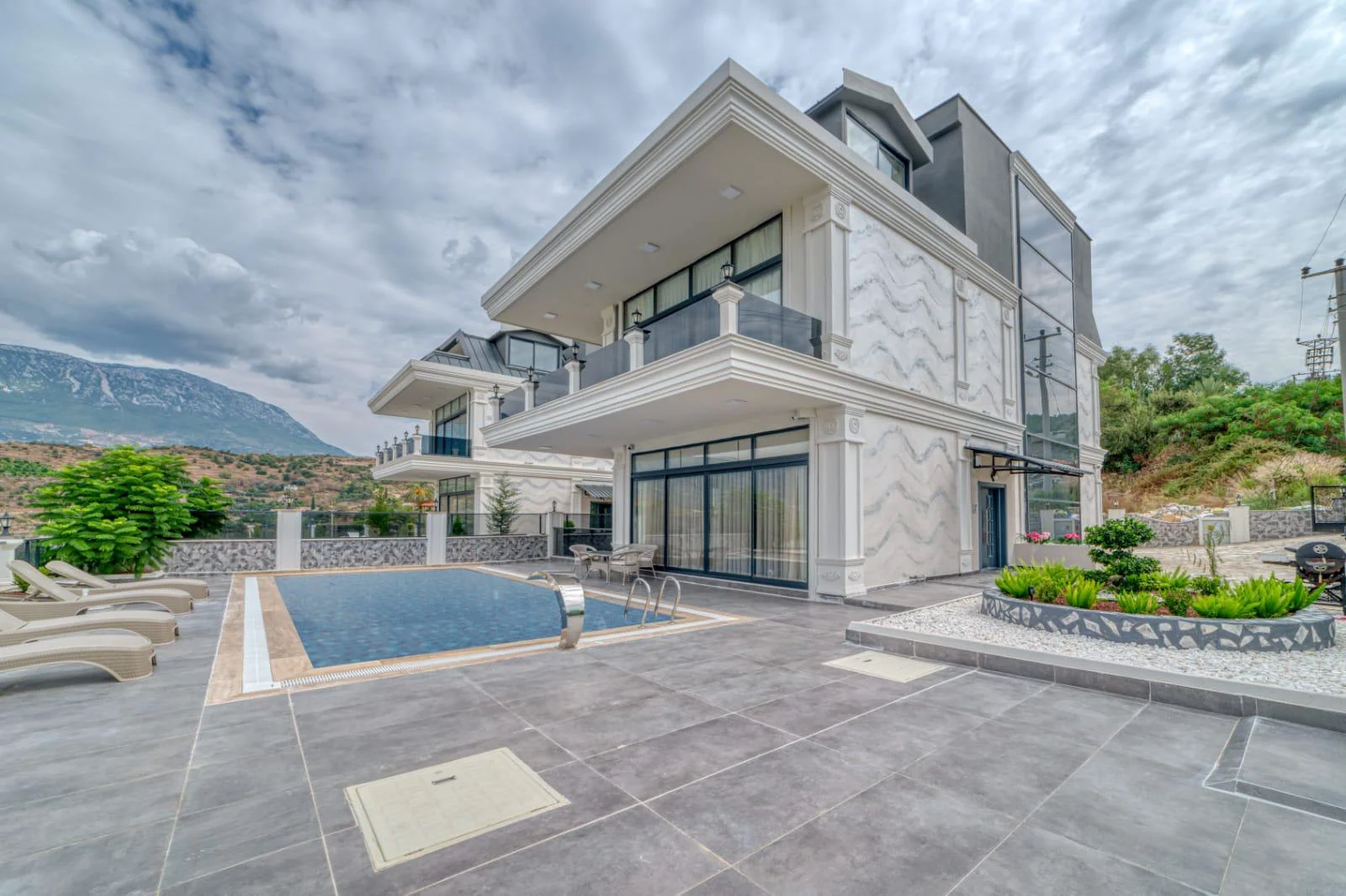 Luxuriöse moderne Villa mit privatem Pool und Bergblick. Außenansicht einer modernen, luxuriösen Villa mit privatem Swimmingpool, gefliester Terrasse mit Liegestühlen und atemberaubendem Bergblick im Hintergrund. Zeitgenössische Architektur.