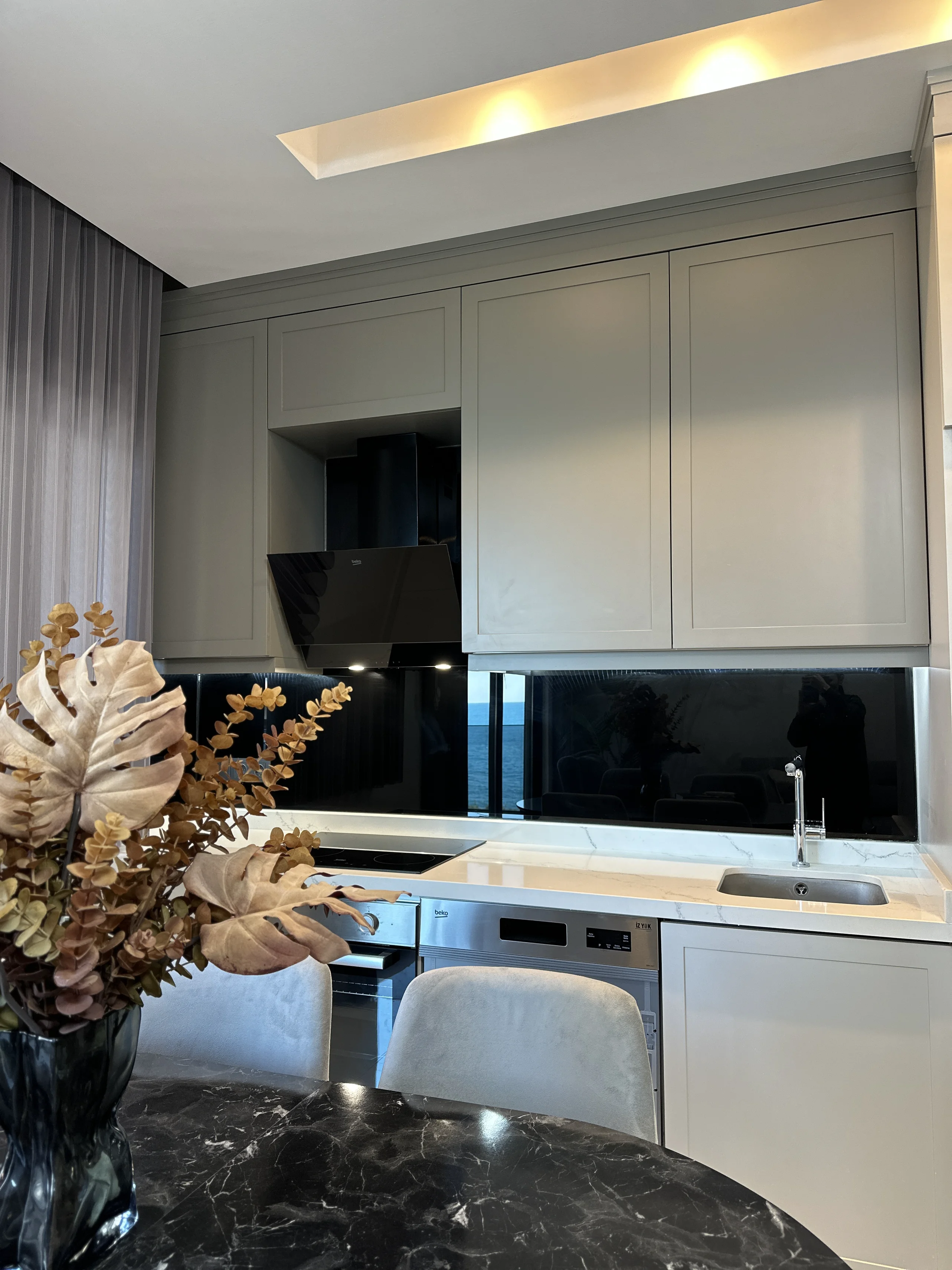 Moderne Küche mit Meerblick und Marmorarbeitsplatten. Eine zeitgenössische Küche mit hellgrauen Schränken, einer weißen Marmorarbeitsplatte und integrierten Geräten. Die schwarze, reflektierende Küchenrückwand bietet einen atemberaubenden Meerblick.