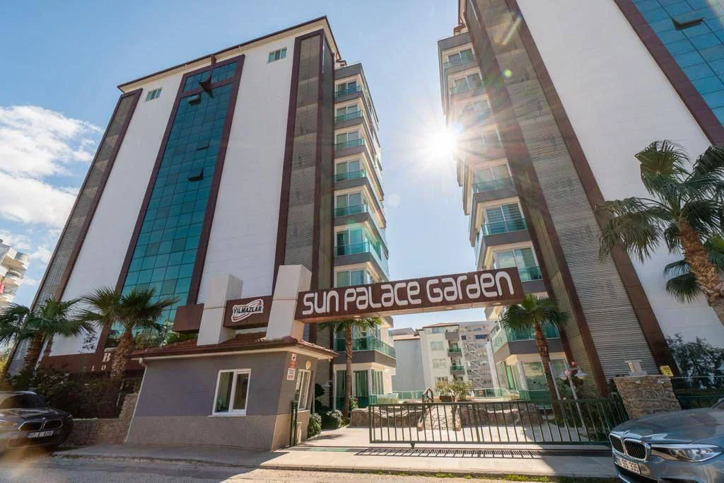 Eingang der modernen Wohnanlage Sun Palace Garden. Außenansicht der modernen Wohnanlage Sun Palace Garden mit zwei hohen Gebäuden, Glasbalkonen, Sicherheitstor und Palmen unter einem sonnigen Himmel in der Türkei.