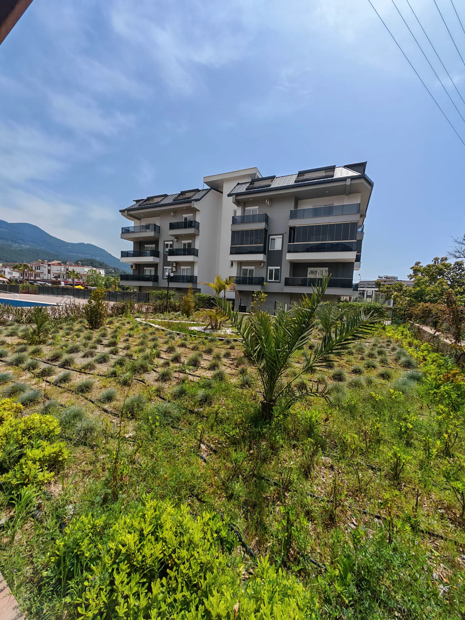 Moderne Außenansicht eines Apartmenthauses mit üppigem grünem Garten. Außenansicht eines modernen Wohngebäudes mit grauer Fassade, Glasbalkonen, einem gepflegten grünen Garten und einem sichtbaren Swimmingpool, mit Bergen in der Ferne.