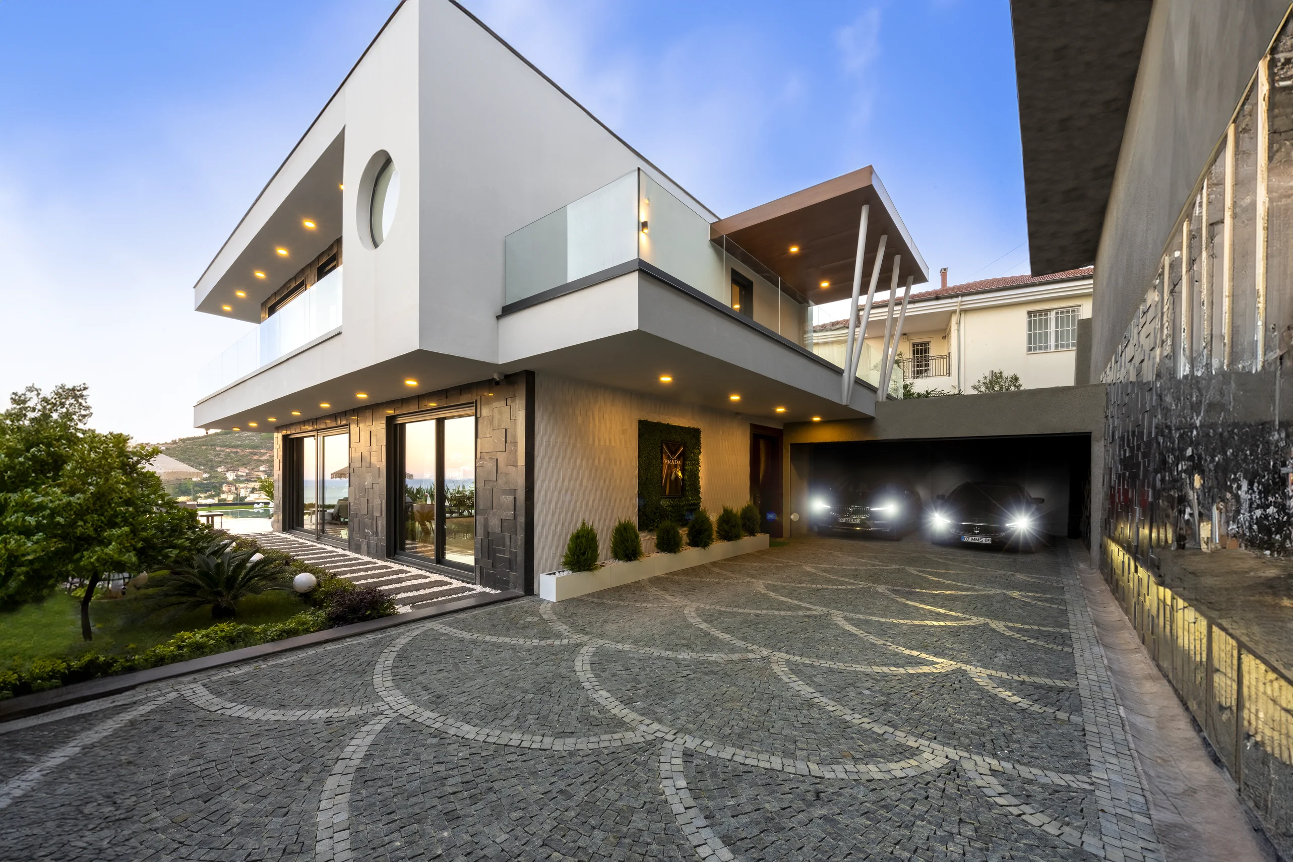 Luxuriöse moderne Villa Außenansicht mit Meerblick und Parkplatz. Außenansicht einer modernen Luxusvilla mit elegantem Design, gemusterter Kopfsteinpflaster-Einfahrt, üppigem Garten, integrierter Garage mit Autos und atemberaubendem Meer- und Bergblick.