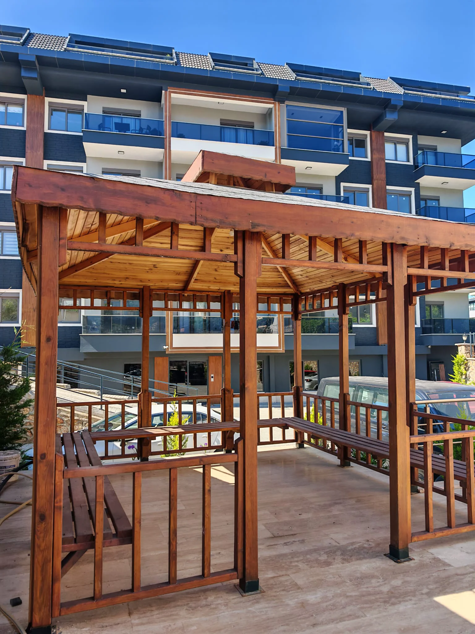 Eleganter Holzpavillon mit Sitzgelegenheiten im Apartmentkomplex. Ein gut gepflegter Holzpavillon mit eingebauten Bänken bietet einen schattigen Sitzbereich im Freien für die Bewohner des modernen Apartmentkomplexes, mit Fliesenboden unter einem klaren blauen Himmel.