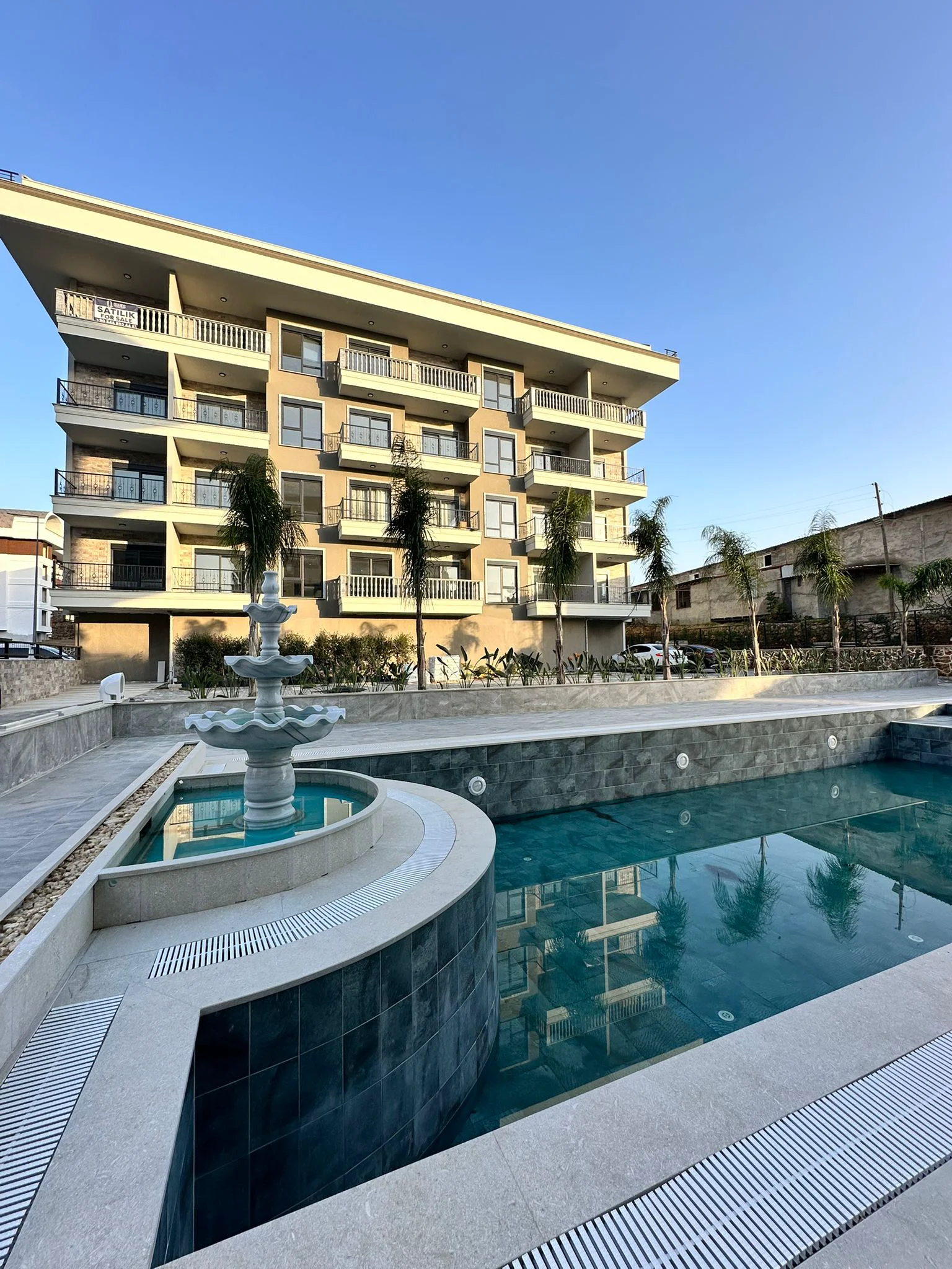 Moderne Wohnanlage mit Außenpool und Springbrunnen. Außenansicht eines modernen mehrstöckigen Wohnhauses mit zahlreichen Balkonen, einem funkelnden blauen Swimmingpool und einem eleganten Etagenbrunnen. Palmen verschönern den gepflegten Außenbereich.