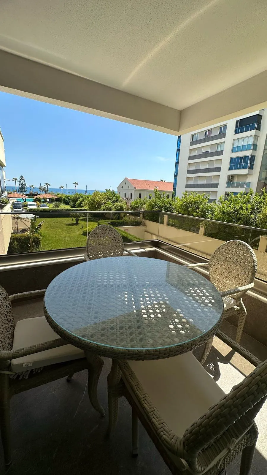 Moderner Balkon mit Meer- und Gartenblick. Moderner Außenbalkon mit einem runden Korbtisch mit Glasplatte und passenden Stühlen. Fliesenboden, Glasgeländer und malerischer Blick auf einen üppig grünen Garten und das funkelnde blaue Meer.